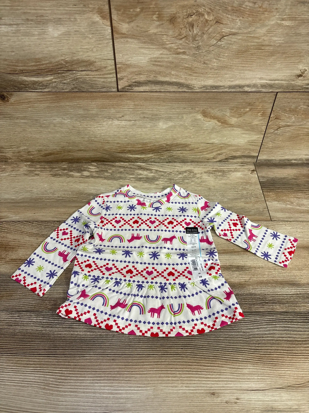 Front view of Okie Dokie Unicorn Fair Isle Peplum Shirt in white, size 6M, showcasing colorful patterned long sleeves on a wood floor