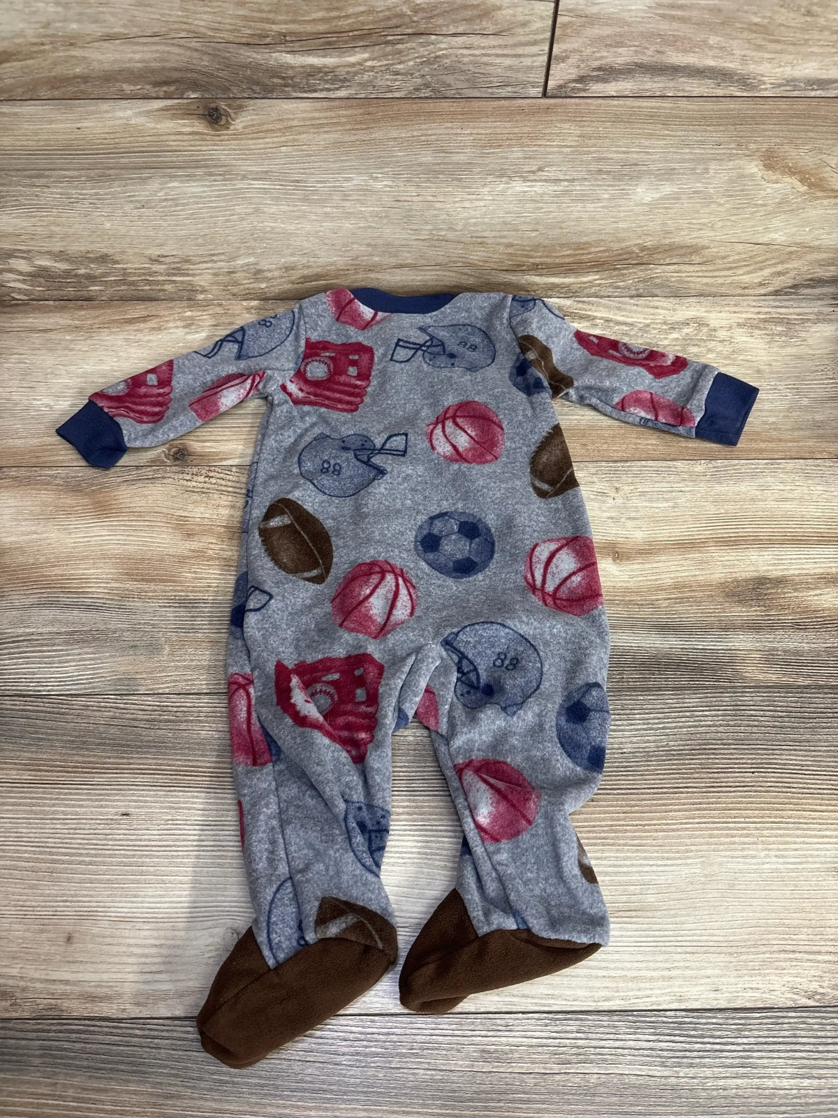 Gray fleece baby sleeper with sports-themed prints including footballs, basketballs, soccer balls, helmets, and gloves on wooden floor