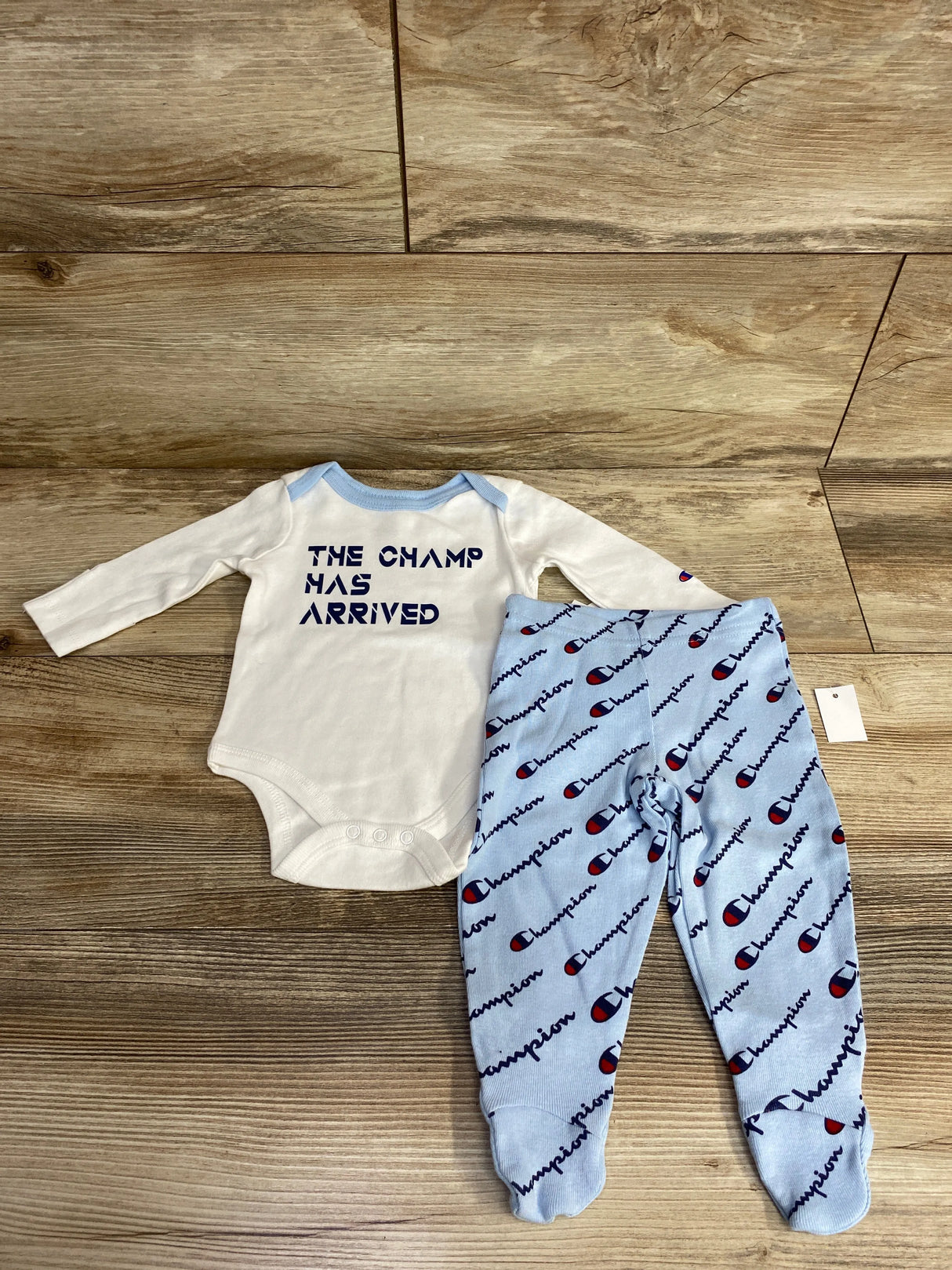 White baby onesie with blue trim reading 'The Champ Has Arrived' and blue Champion logo footed pants on wooden floor
