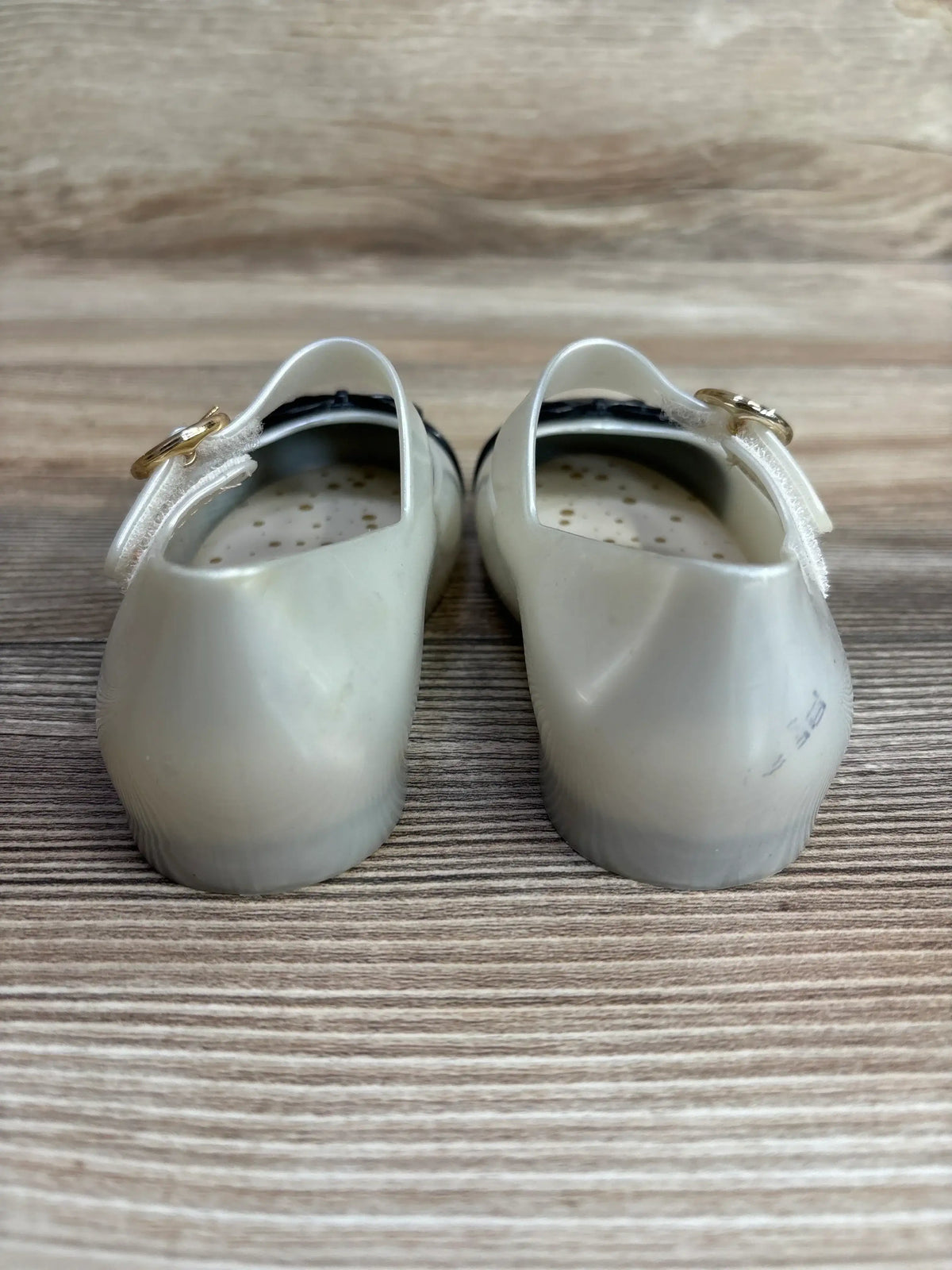 Back view of white translucent toddler shoes with perforated inner soles and gold buckle straps on wood surface
