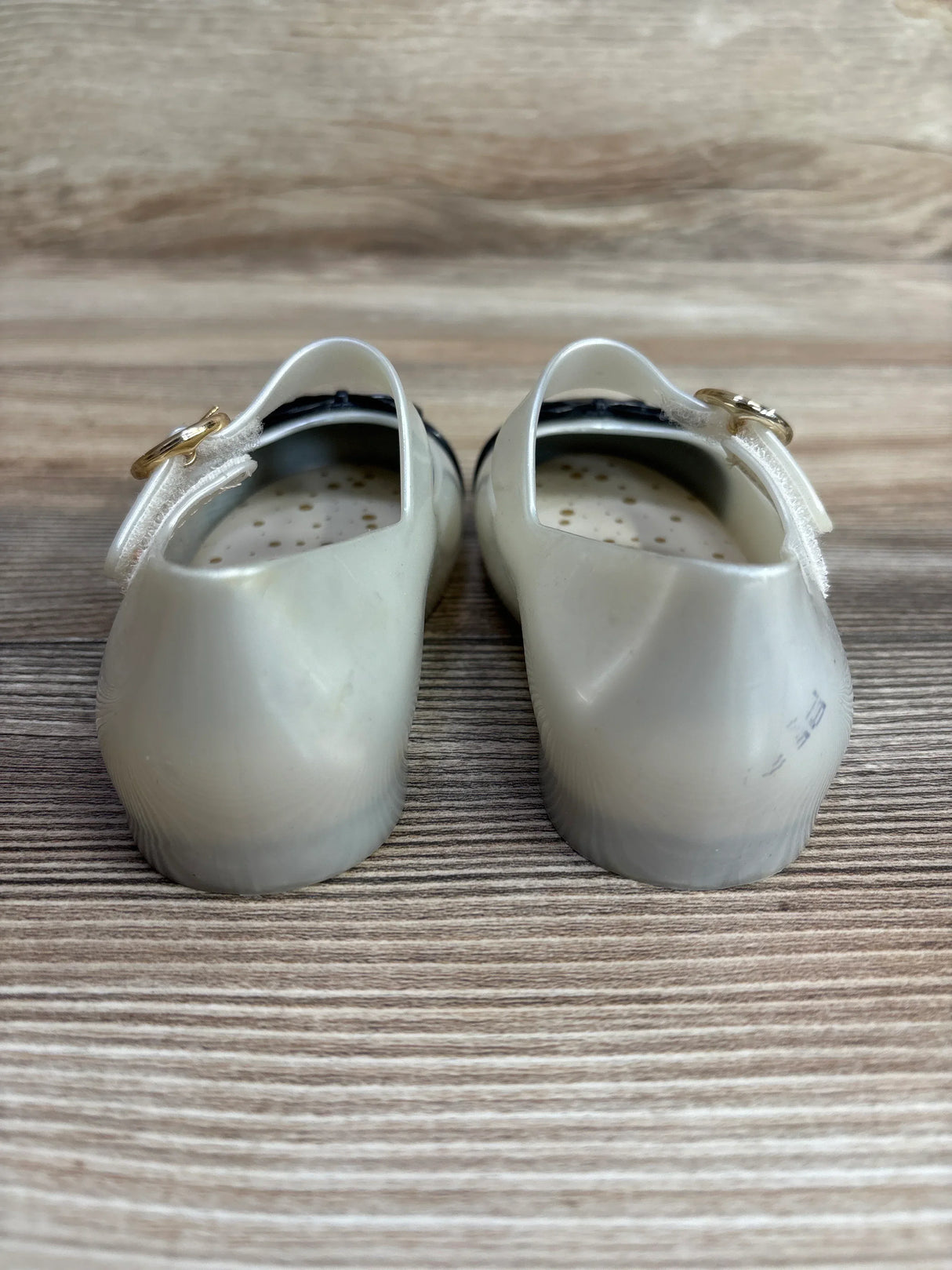 Back view of white translucent toddler shoes with perforated inner soles and gold buckle straps on wood surface