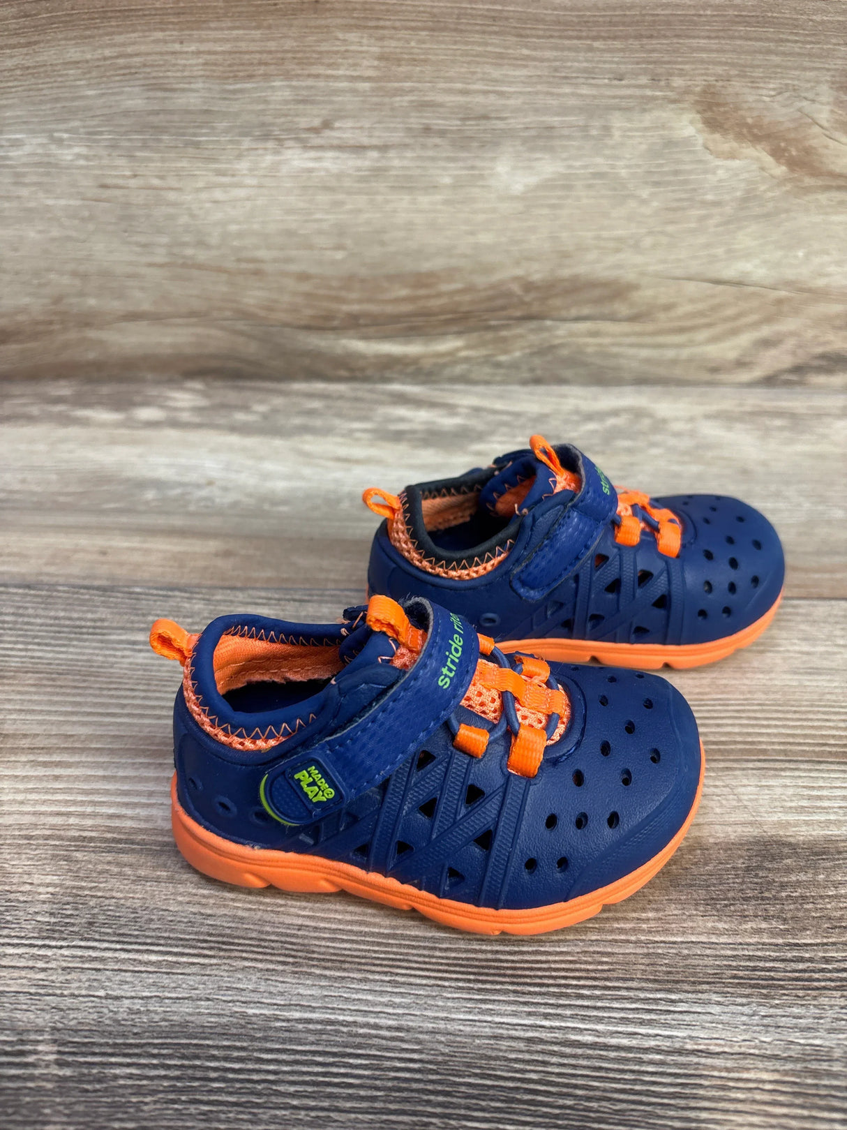 Side view of Stride Rite Phibian sneakers with navy perforated upper, orange sole, Velcro strap on wood background
