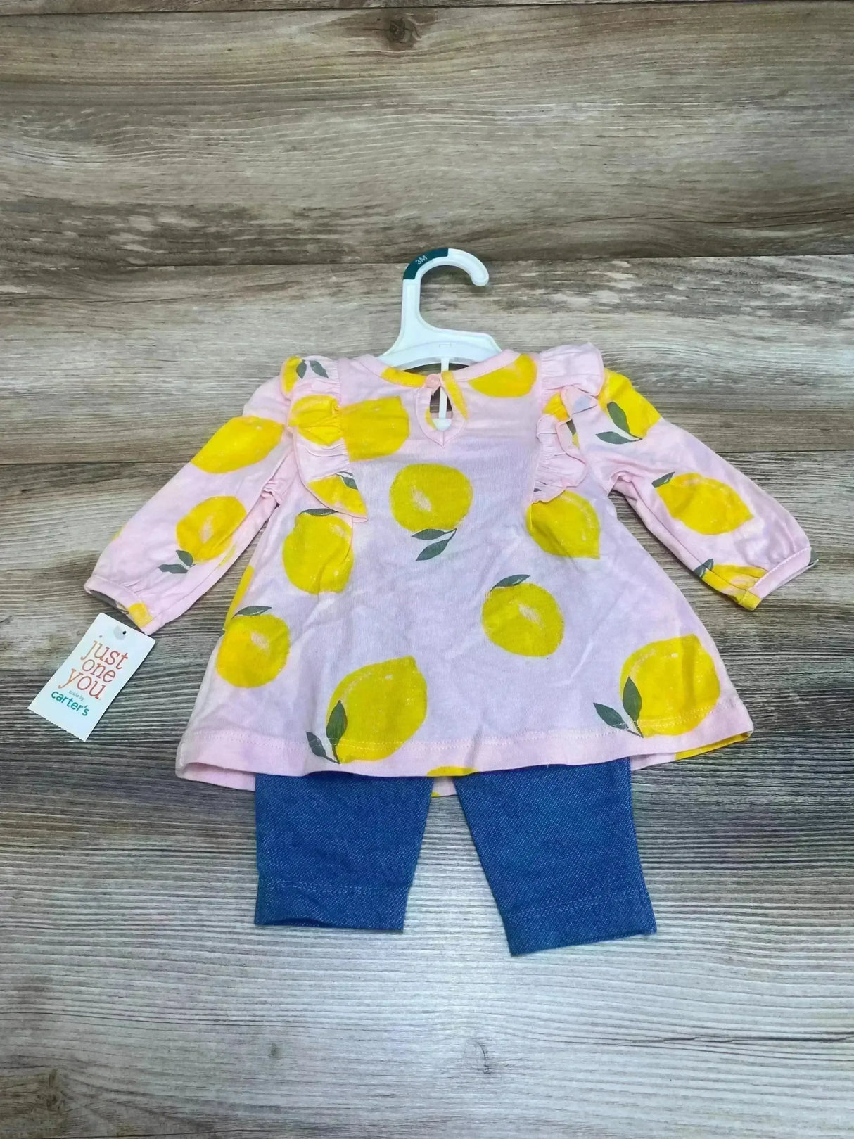 Flat-lay of infant two-piece set showing a pink long-sleeve lemon-print top with ruffle shoulders and keyhole tie, paired with blue leggings on a wood background