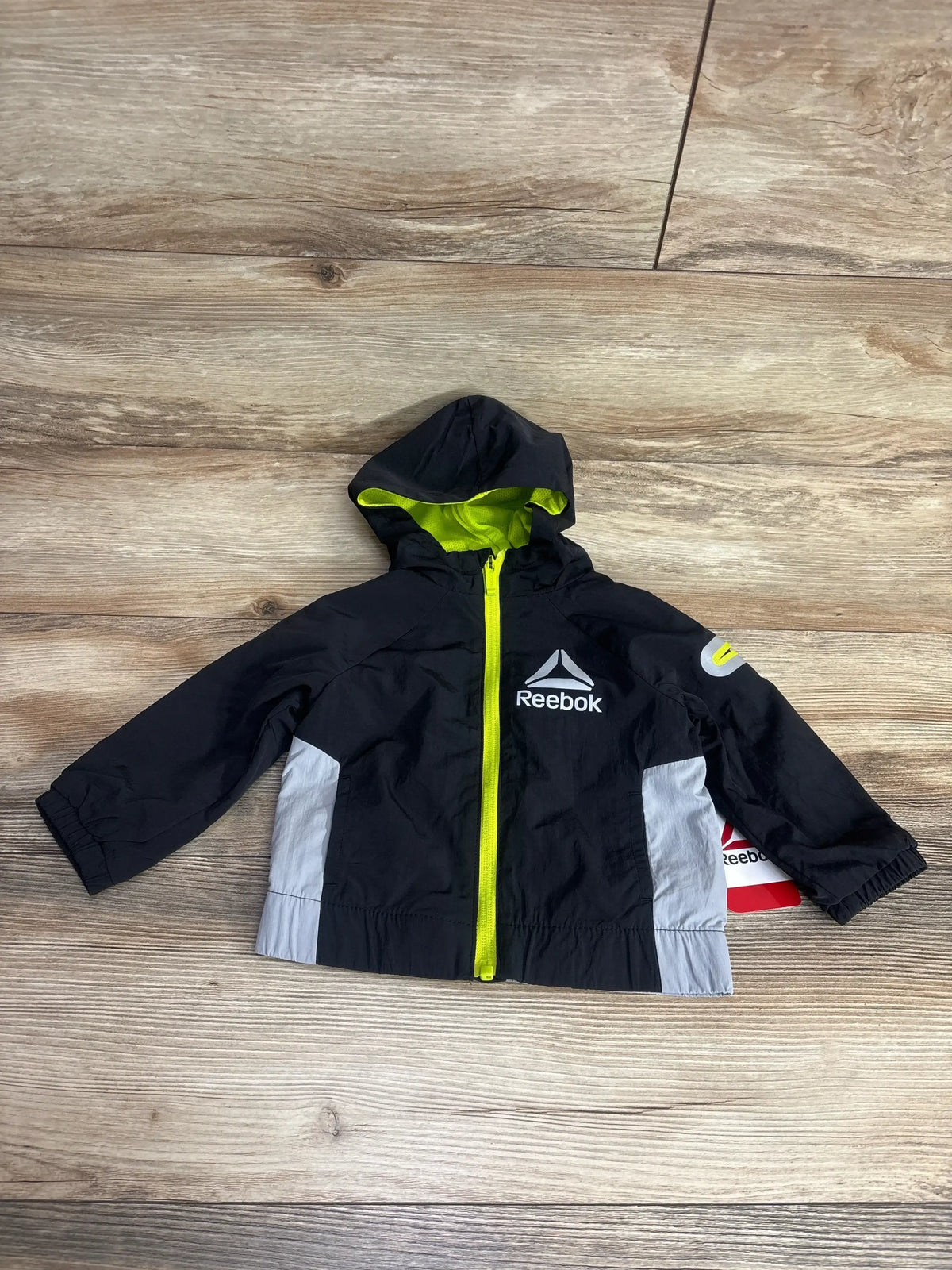 Black and gray Reebok toddler windbreaker with hood and neon yellow zipper, laid flat on wood floor