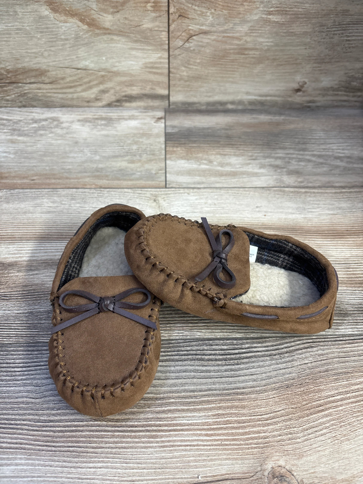Top and side view of brown suede moccasin slippers with faux-fur lining and bow detail on wood floor
