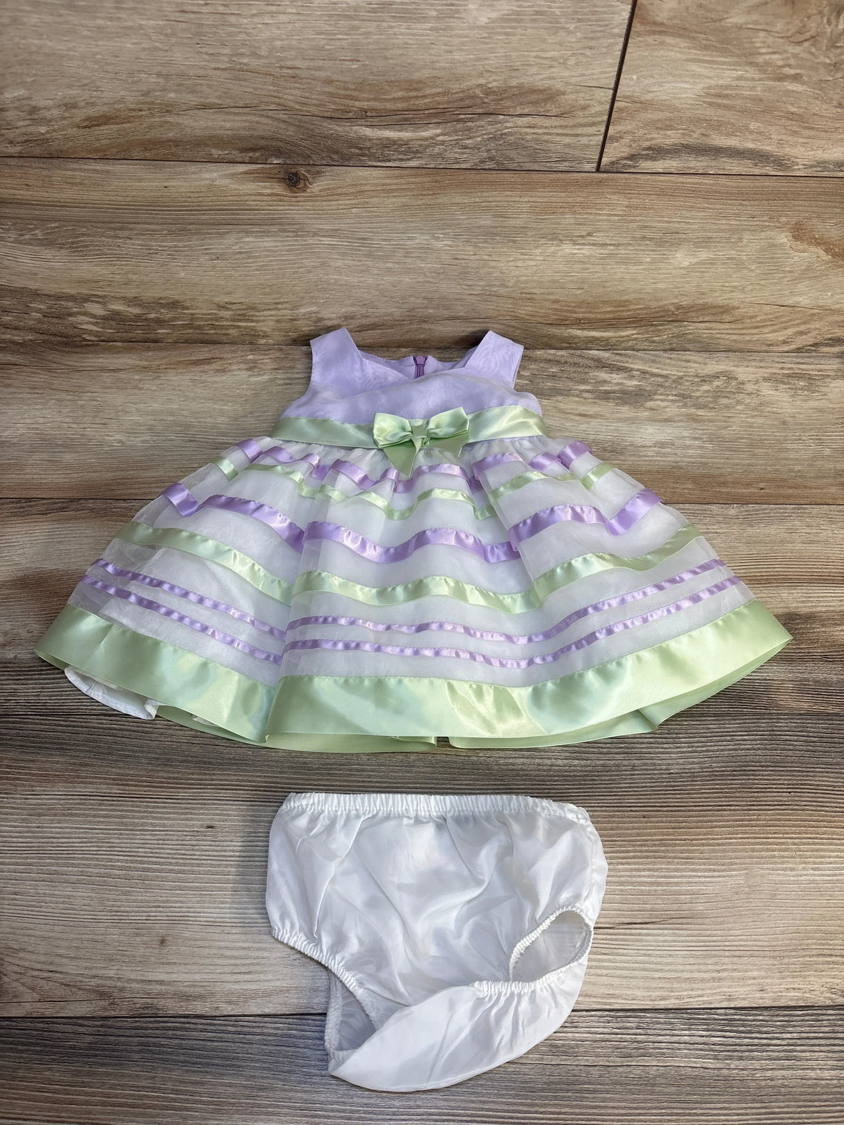 Flat lay of Bonnie Baby 2pc sleeveless dress showcasing lilac and green ribbon striped skirt with bow and white bloomers on wood plank floor