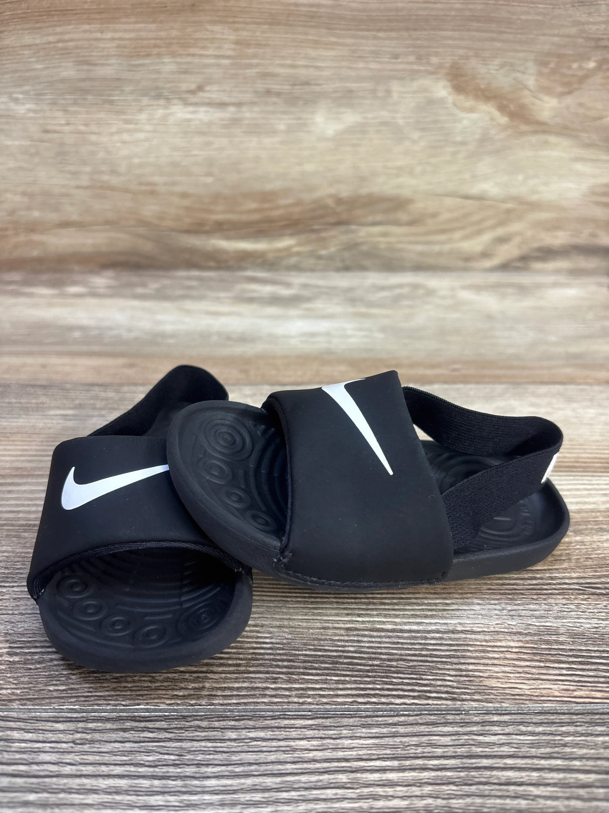 Angled top view of black Nike Kawa toddler slide sandals showing white Swoosh logo, elastic heel strap on wood surface