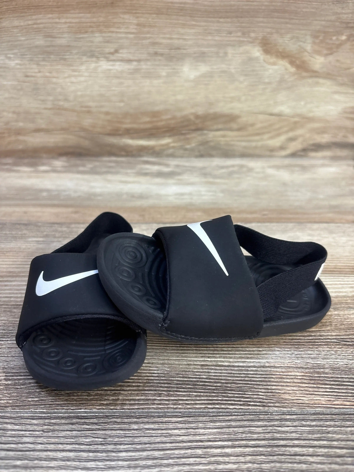 Angled top view of black Nike Kawa toddler slide sandals showing white Swoosh logo, elastic heel strap on wood surface