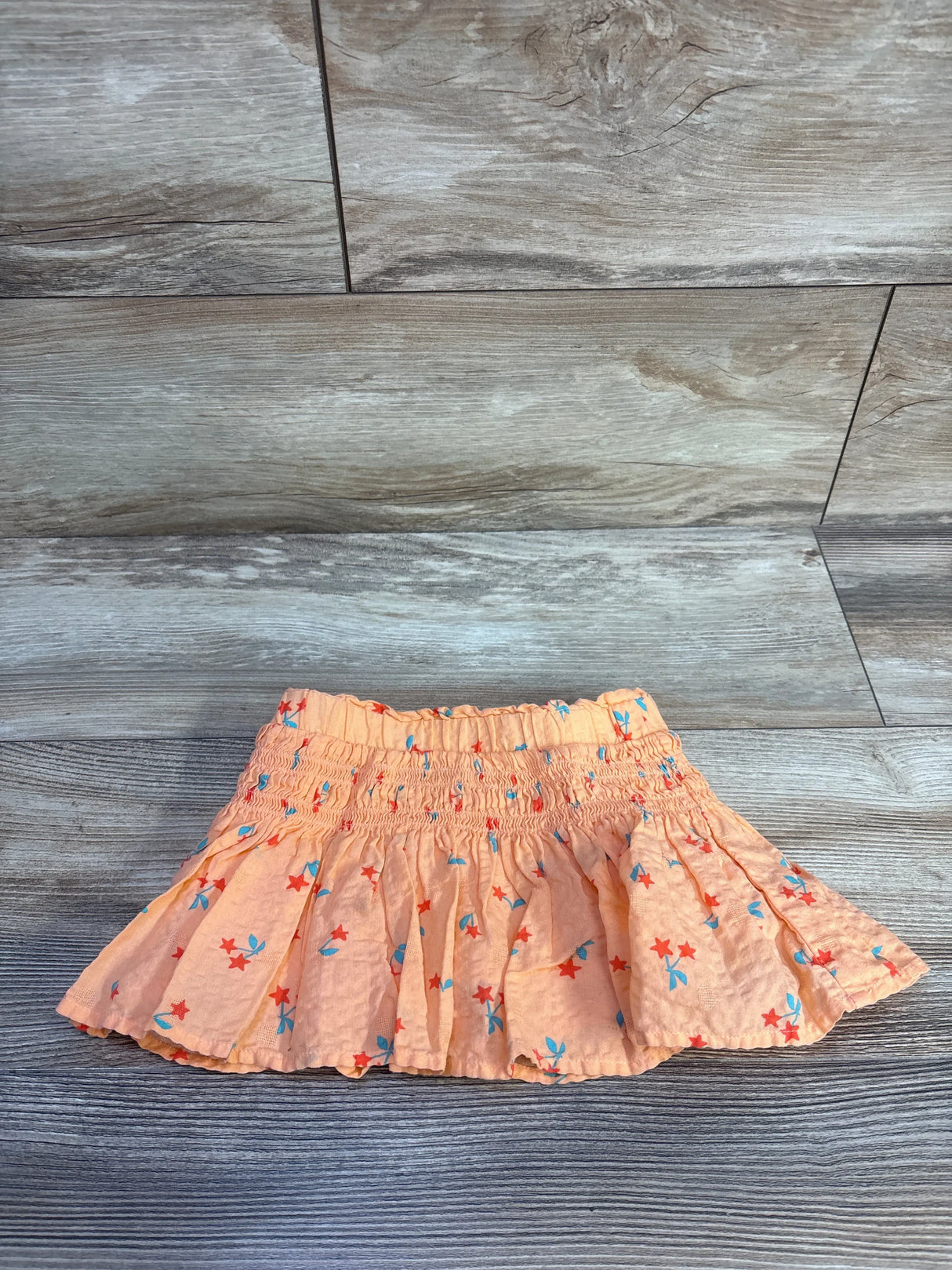 Top-down view of Cat & Jack smocked toddler skort showcasing gathered smocked waistband and red star with blue bow print on wood plank background, size 18 months