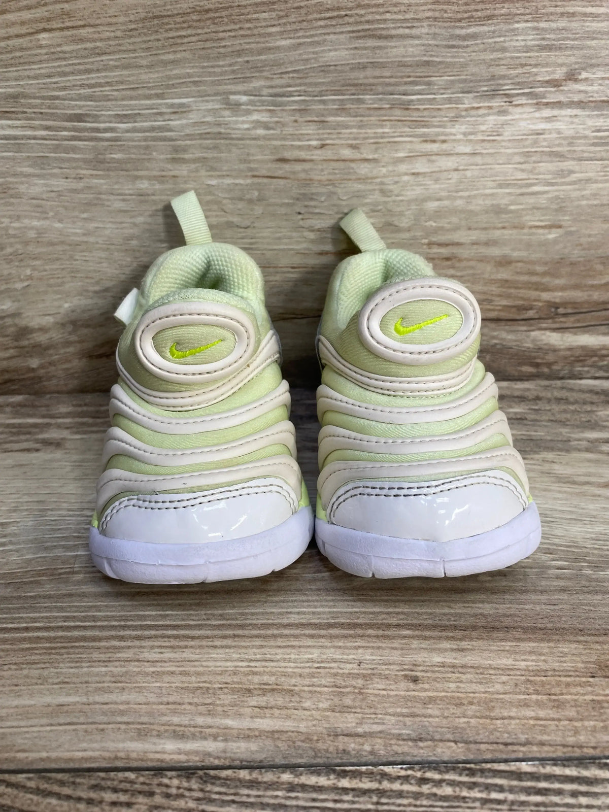 Front view of toddler Nike Dynamo Free sneakers in pale lime green and white showing layered ribbed upper and pull tabs on woodgrain background