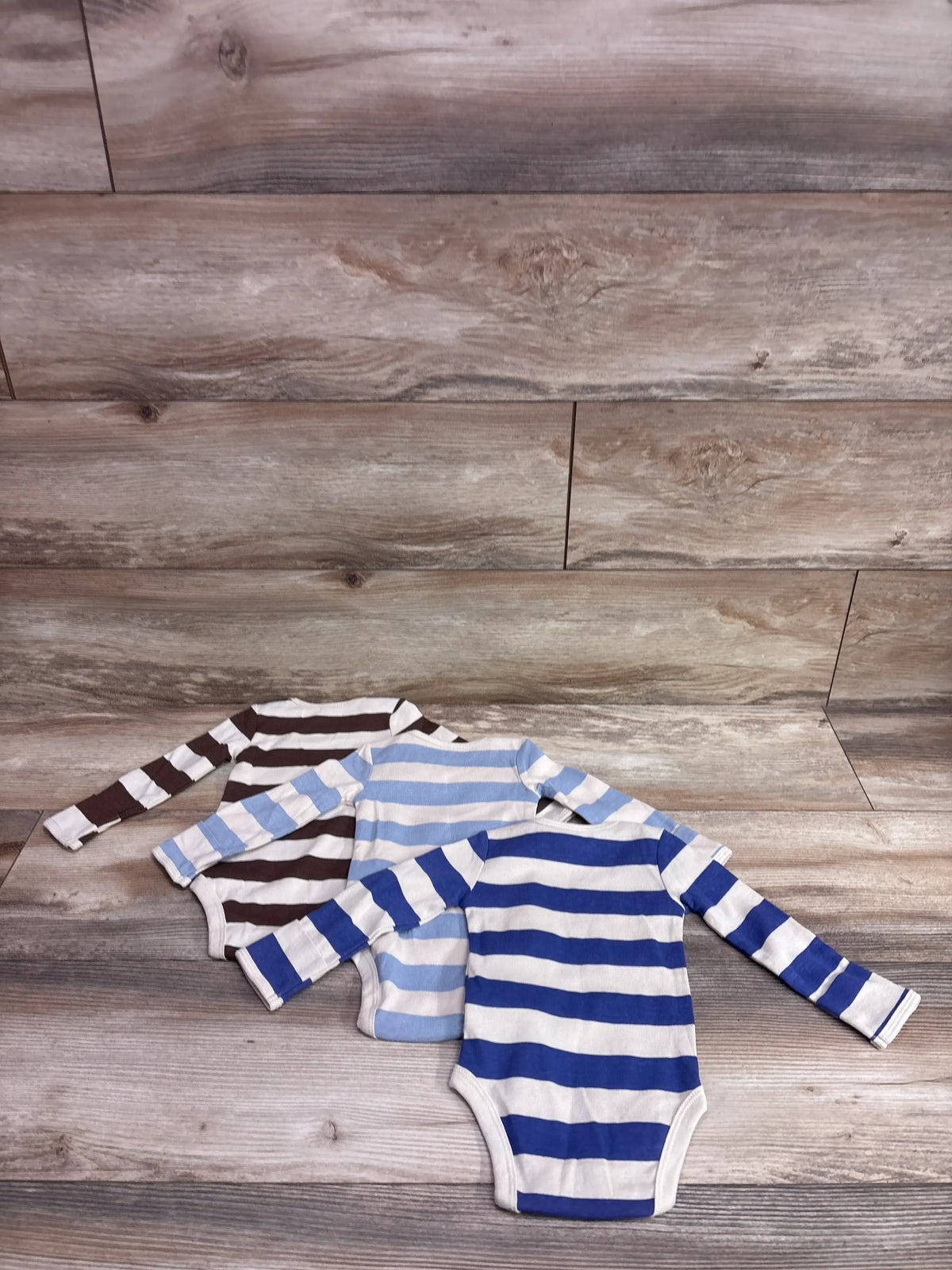 Front view of Carter’s three long sleeve striped bodysuits in brown, light blue, and royal blue on a wood plank background
