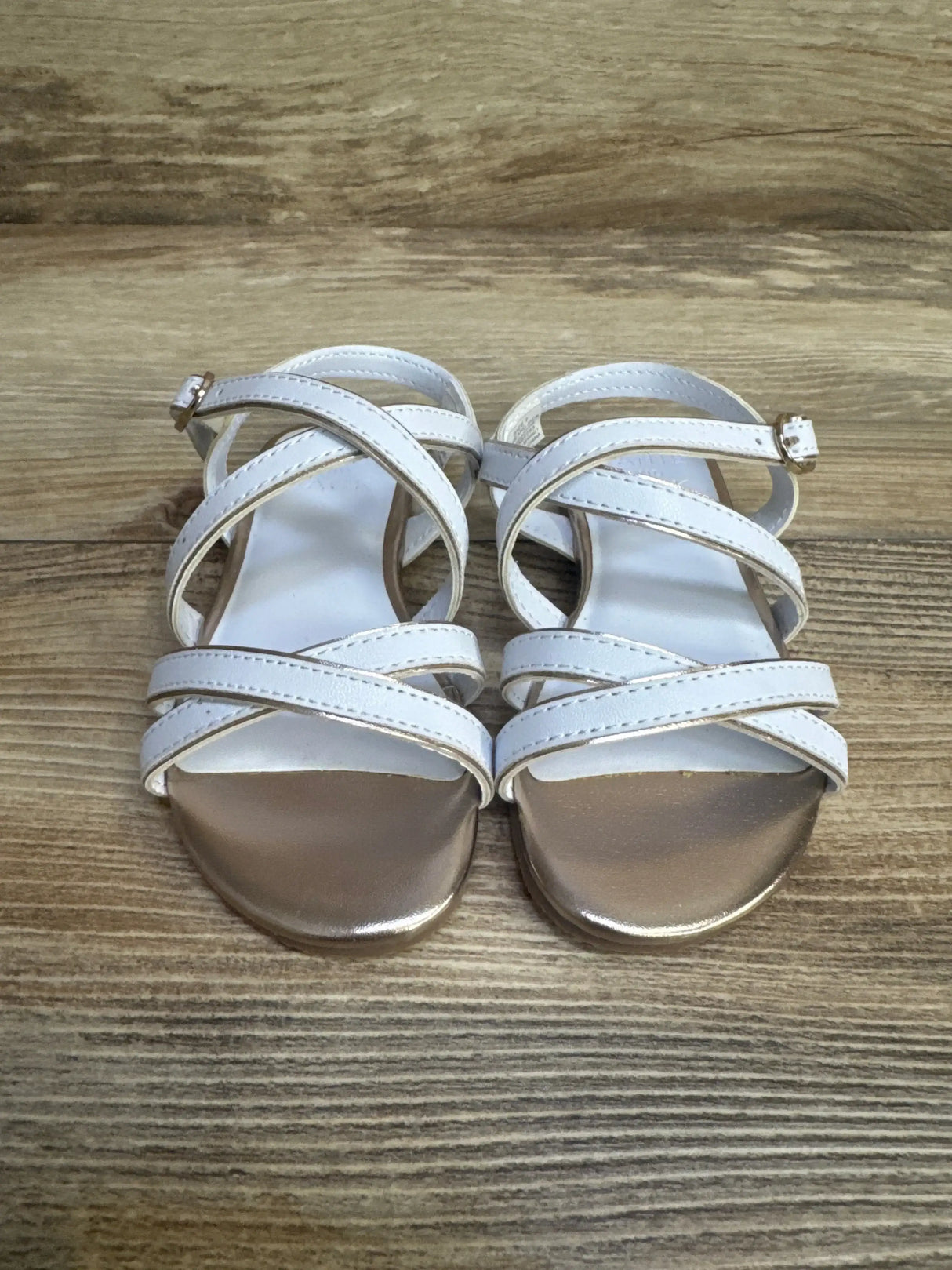 Top-down view of white Janie & Jack toddler sandals showing metallic gold toe trim and crisscross straps on wood floor