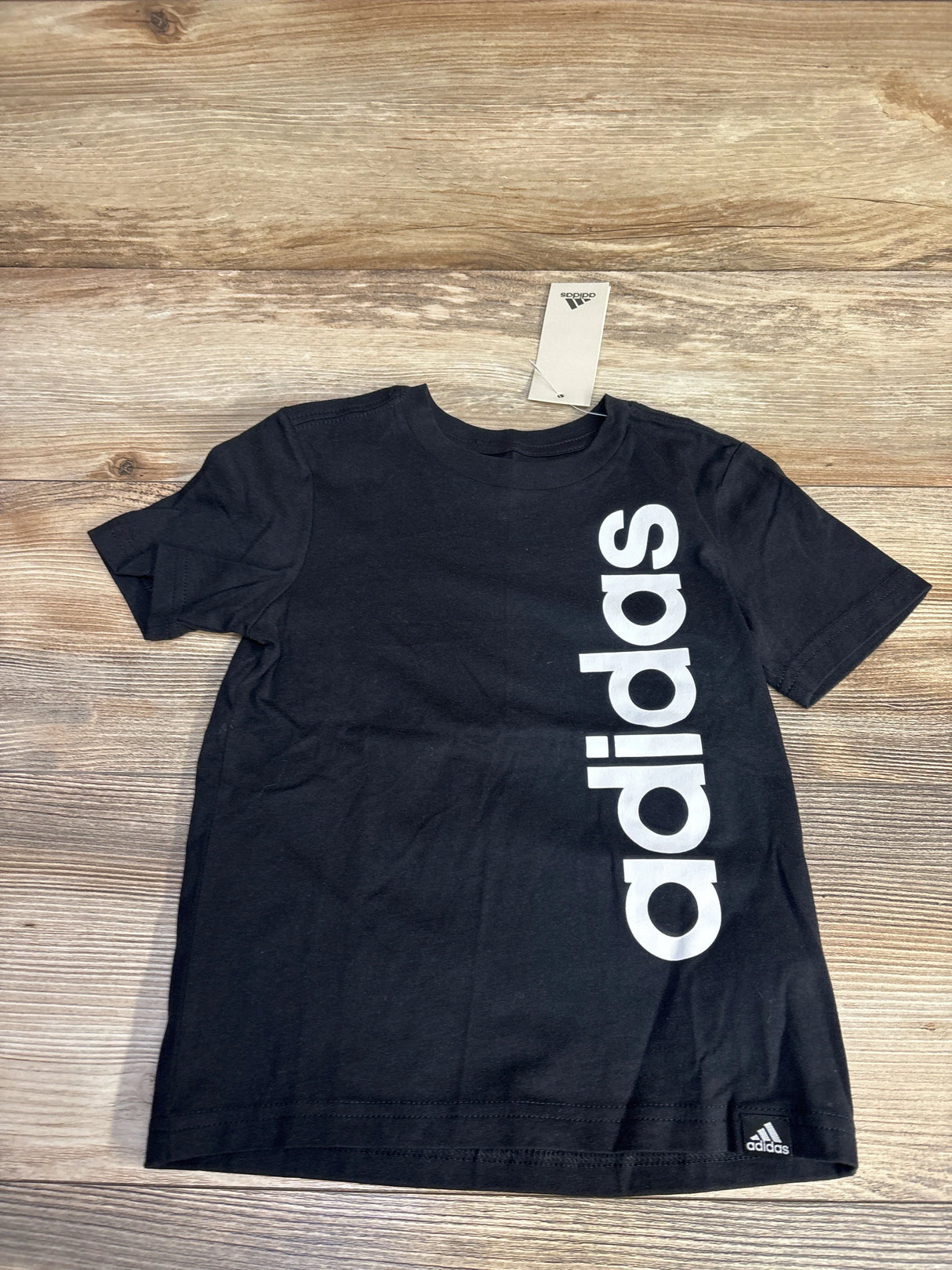 Black Adidas t-shirt with large vertical white logo on wooden surface and Adidas tag attached