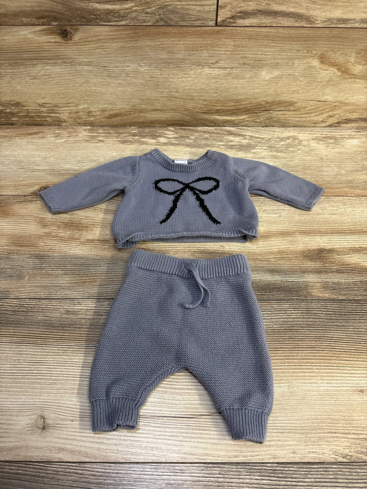 Flat lay of newborn blue knit shirt and pants set showcasing black bow on sweater, drawstring waist and ribbed cuffs on wood background