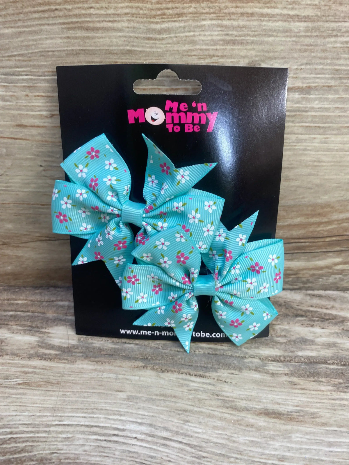 Front view of two teal floral fanned out hair bows clipped to a black Me n Mommy To Be card on a wood grain surface