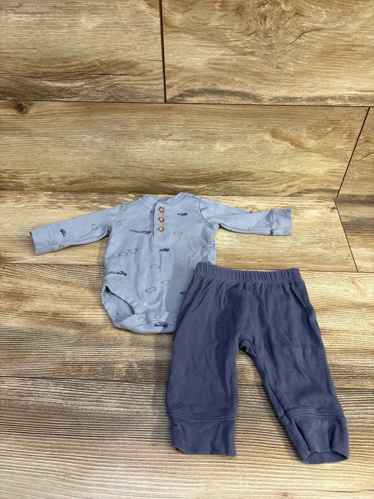 Overhead of newborn 2-piece: light-blue airplane-print bodysuit with wooden buttons and navy elastic pants on wood