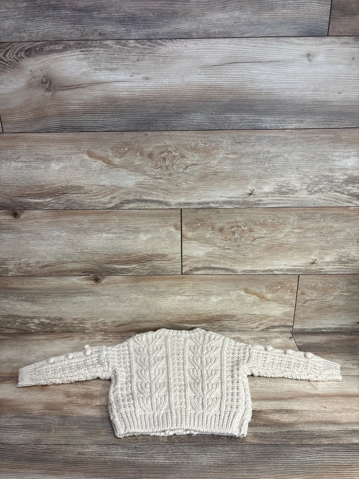 Cable knit cream baby sweater with long sleeves laid flat on wood background