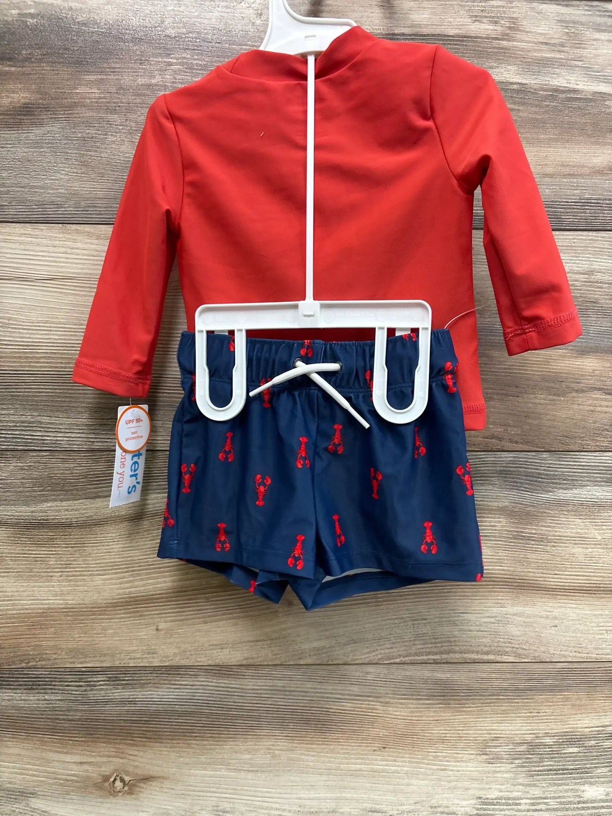 Back view of baby 2-piece rash guard set with red long-sleeve top and navy swim trunks with red lobster print on wood