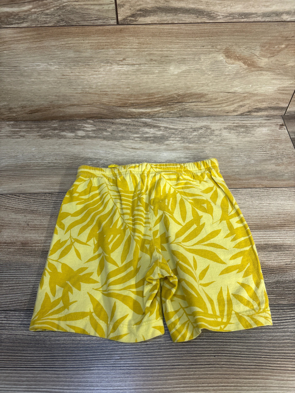 Yellow kids shorts with leaf print, elastic waistband, laid flat on wood background