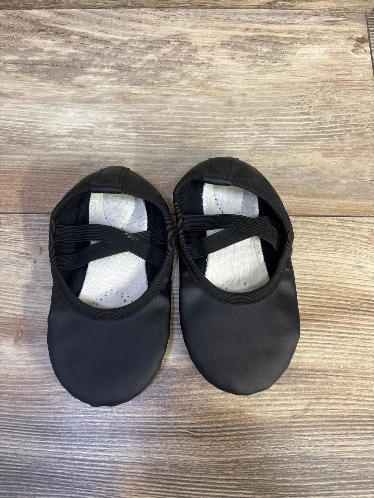 Top-down view of black baby ballet shoes showcasing crossed elastic straps and white insoles on wood floor
