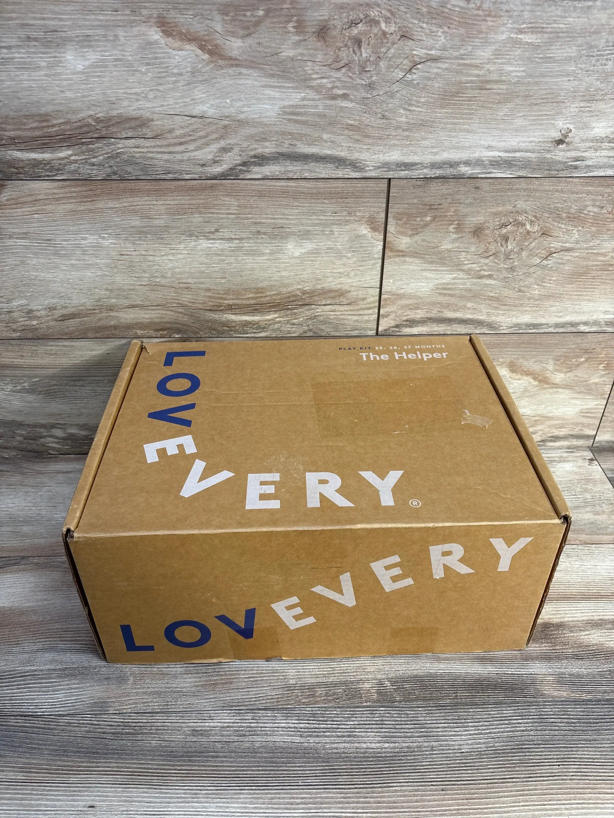 Three quarter view of Lovevery The Helper Play Kit kraft box with white and blue Lovevery branding on wood plank floor