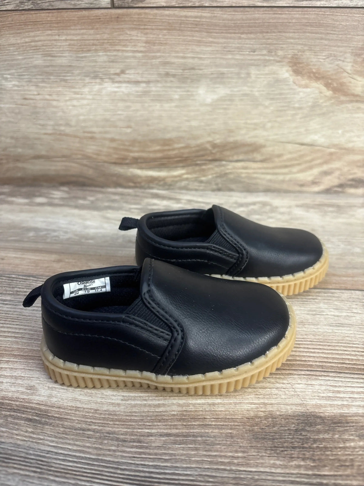 Three-quarter view of OshKosh Victor slip-on sneakers in black, size 4C, showcasing tan ribbed soles on a wood surface