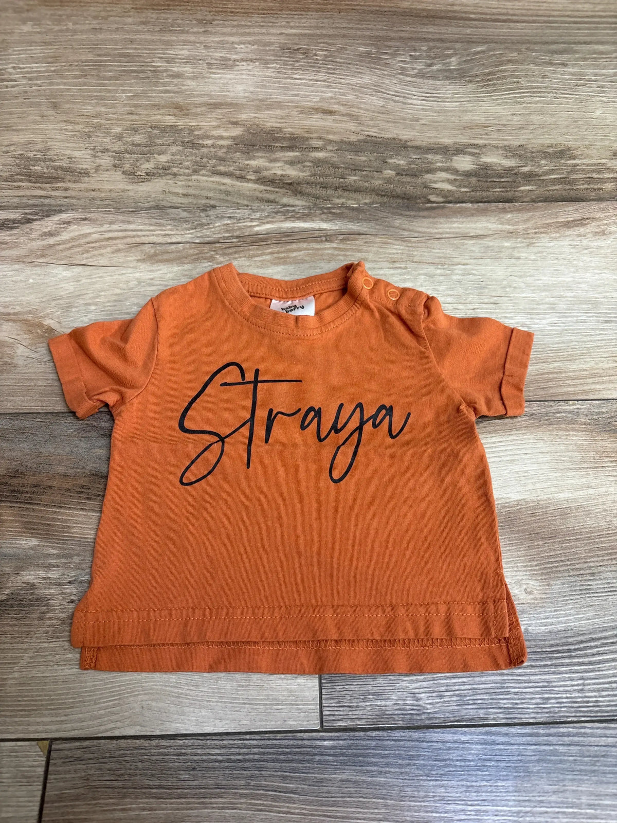Orange baby t-shirt with 'Straya' text on wood floor background