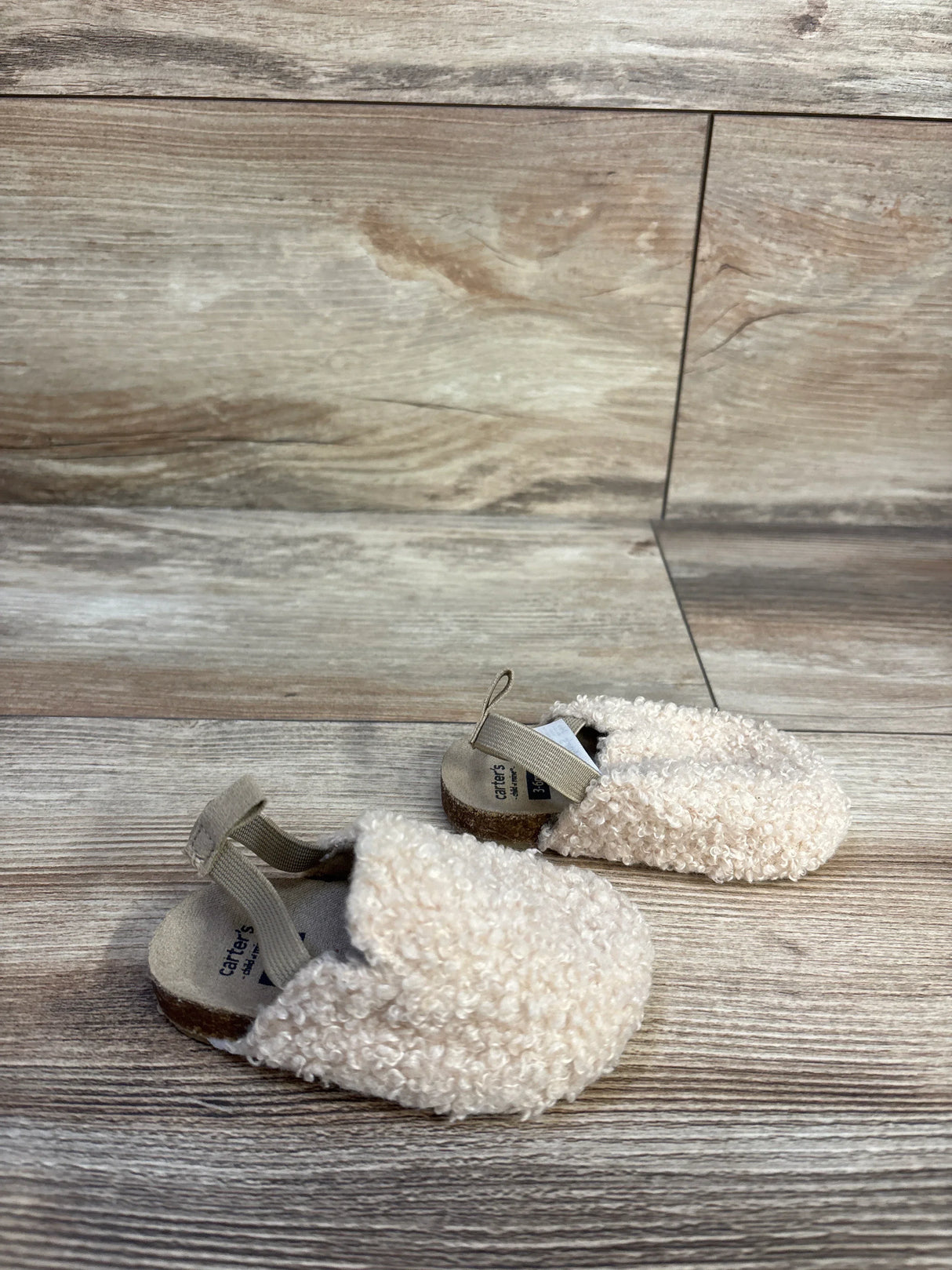 Angled view of Child of Mine slip-on baby clog size 2C showing beige curly faux-sherpa upper and elastic heel strap on light wood background