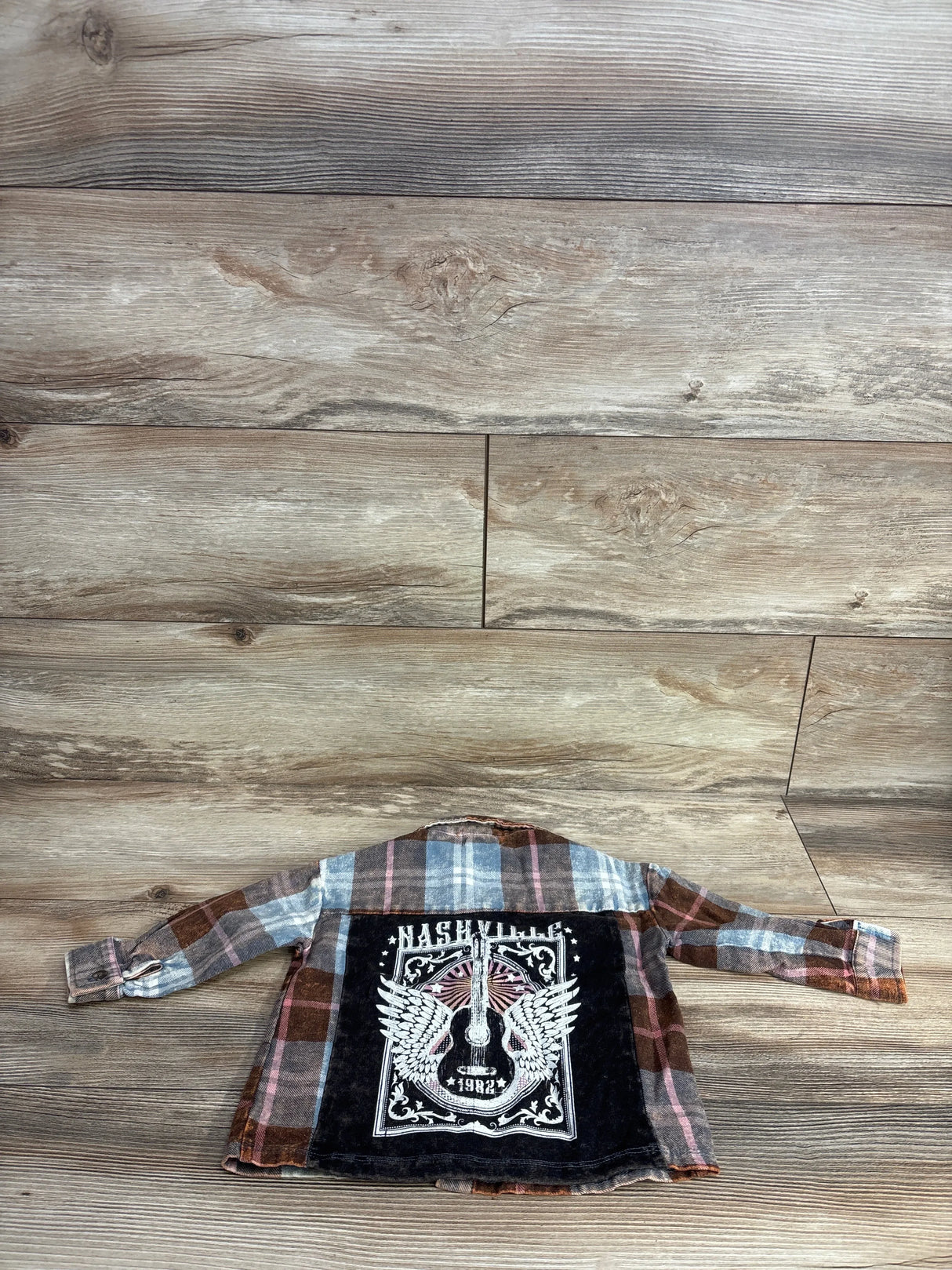 Child's plaid button-up shirt with Nashville 1982 winged guitar graphic on back, laid flat on wooden floor