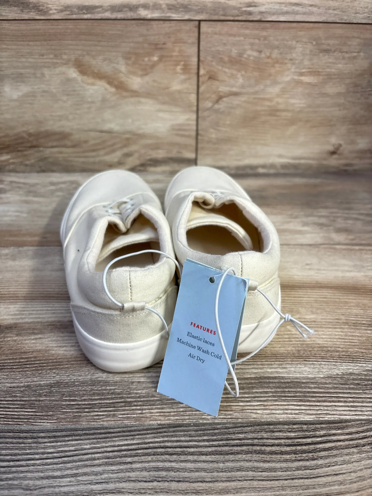 Rear view of off-white Tyler slip-on toddler sneakers showing elastic laces and attached tag on wood background
