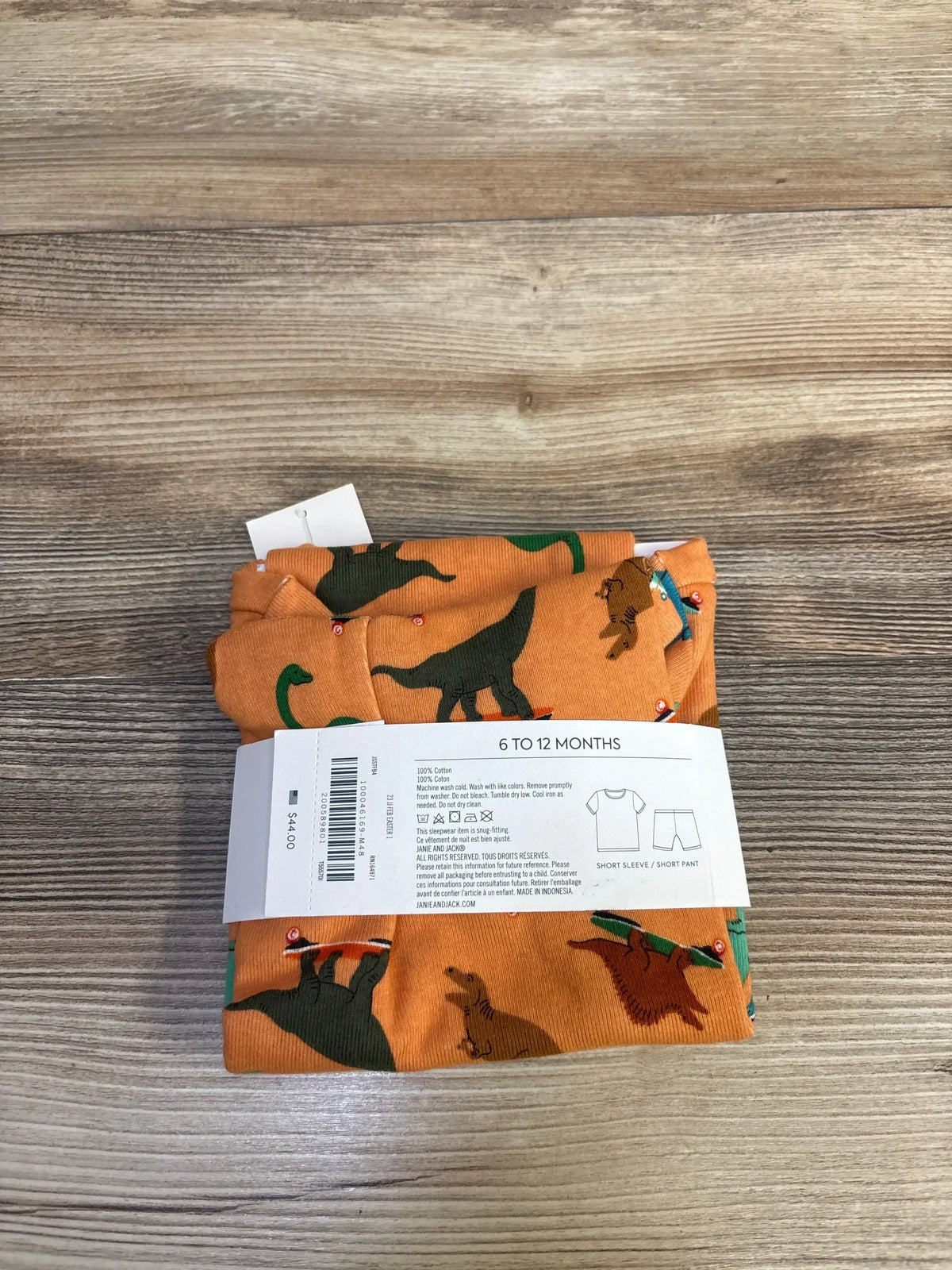 Top-down view of folded Janie & Jack 2-piece baby pajama set showcasing orange fabric with skate-riding dinosaur print and visible size label on wood surface