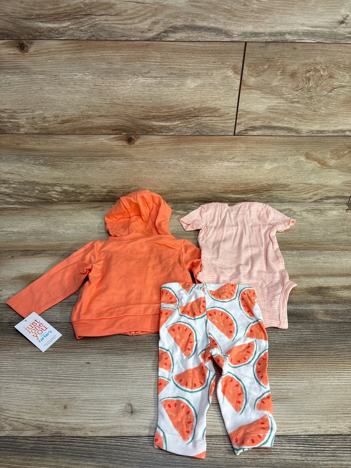 Flat lay of 3-piece watermelon set with peach hooded zip jacket, striped pink bodysuit, and watermelon leggings on wood floor
