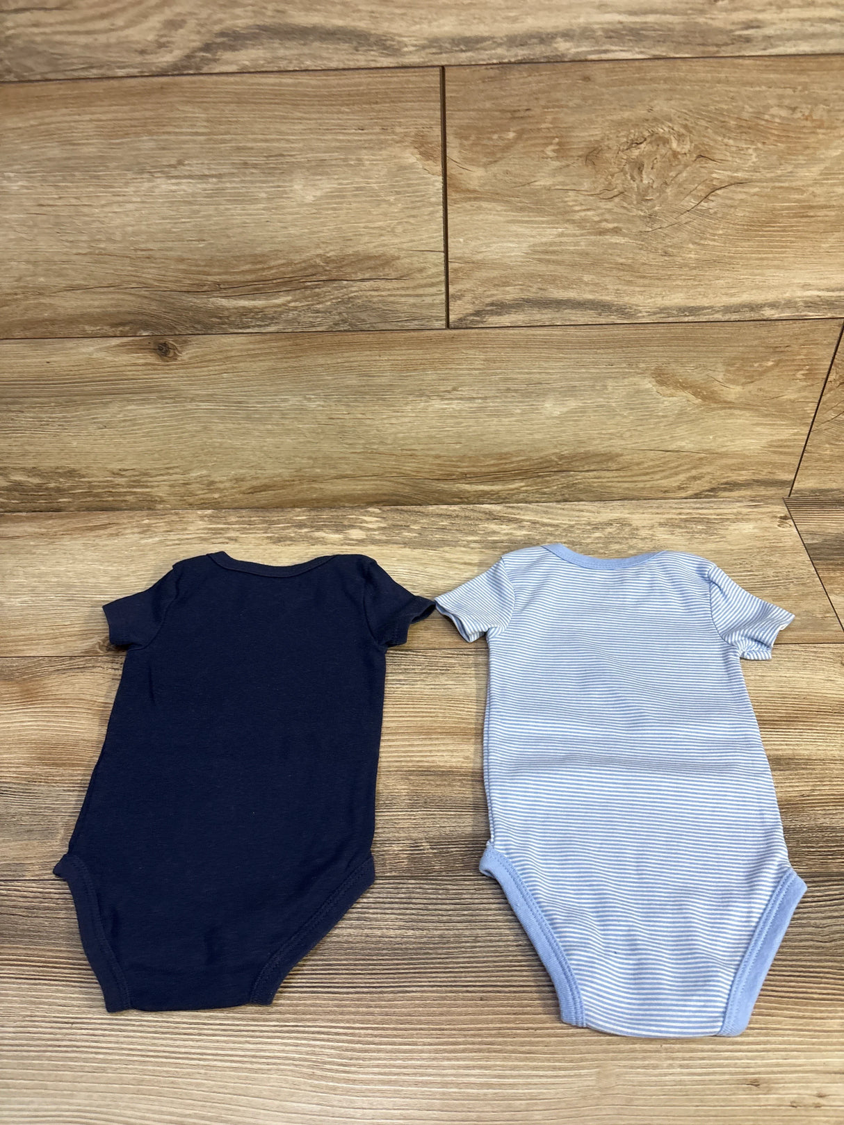 Flat lay of two Nike baby bodysuits (2-pack), solid navy and light-blue stripe, size 9M, on wood background