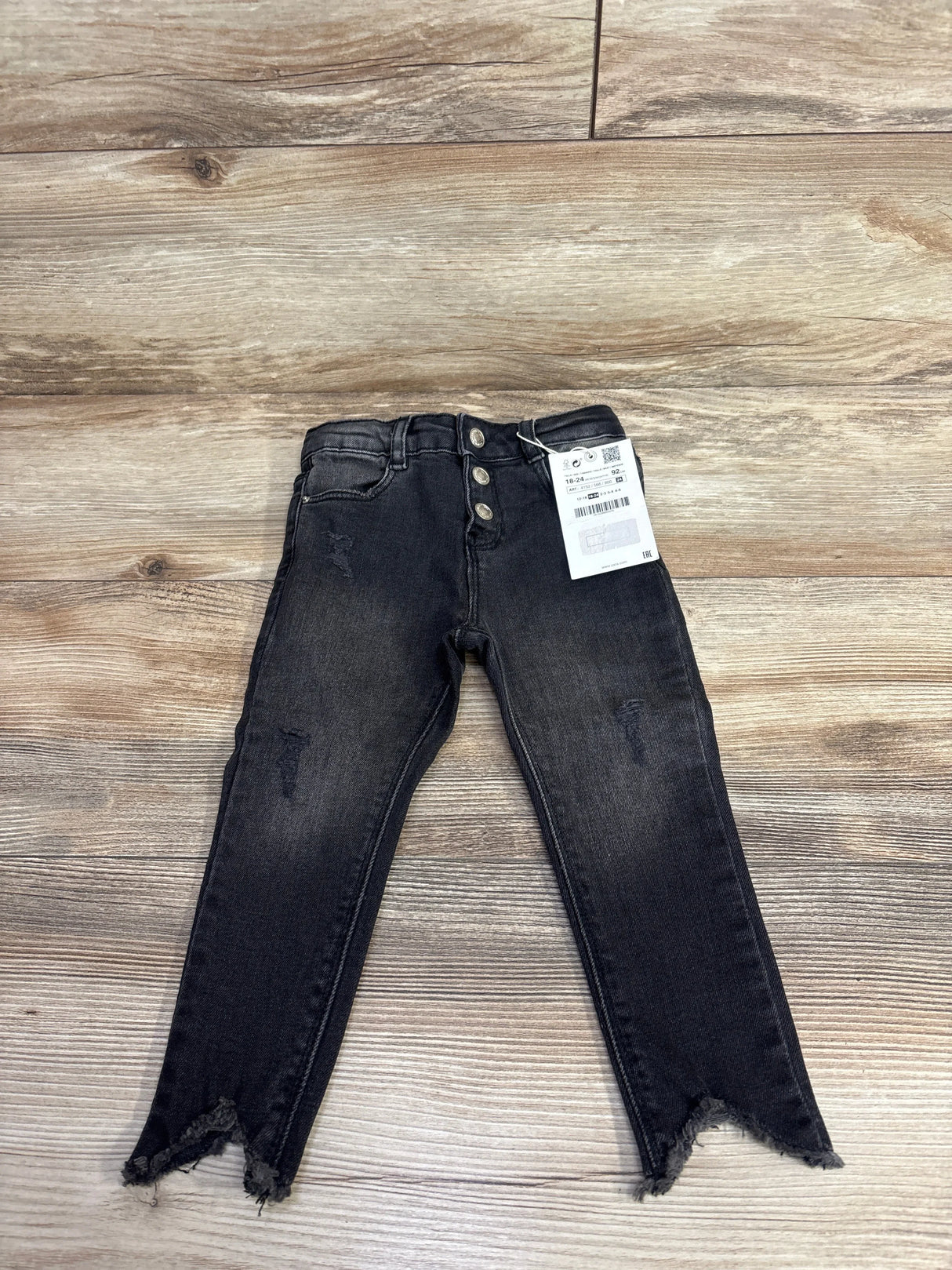 Black distressed kids jeans with three buttons and frayed hems laid flat on wooden surface