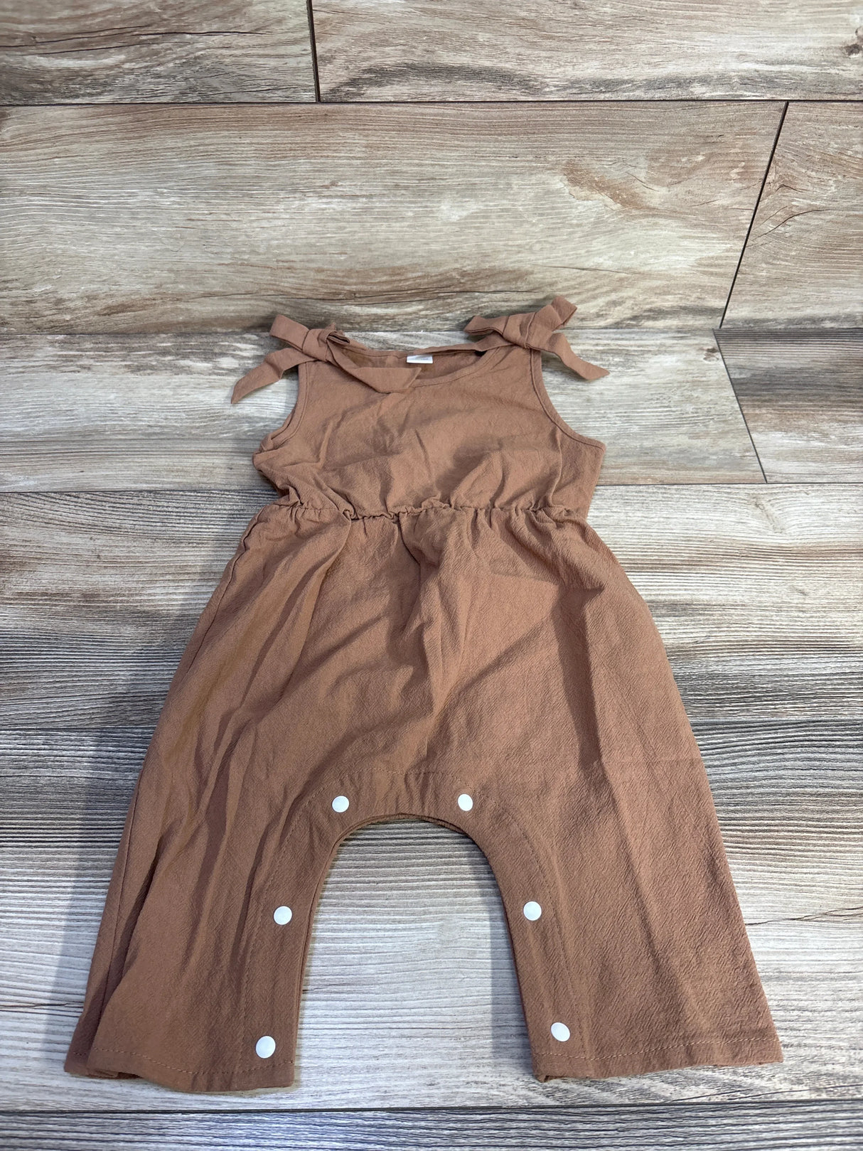 Flat lay of brown sleeveless baby jumpsuit with tied shoulder bows and snap-crotch closure on wood plank background