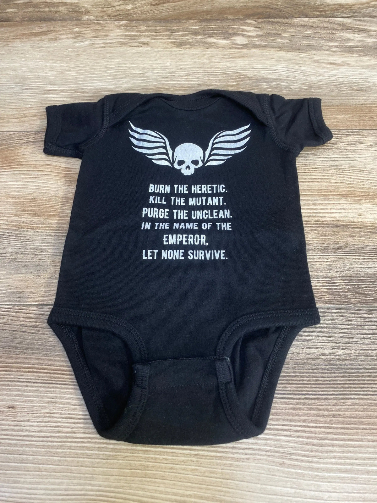 Front flat lay of black Rabbit Skins baby bodysuit with white skull-and-wings and printed slogan 'Burn the heretic' on wood