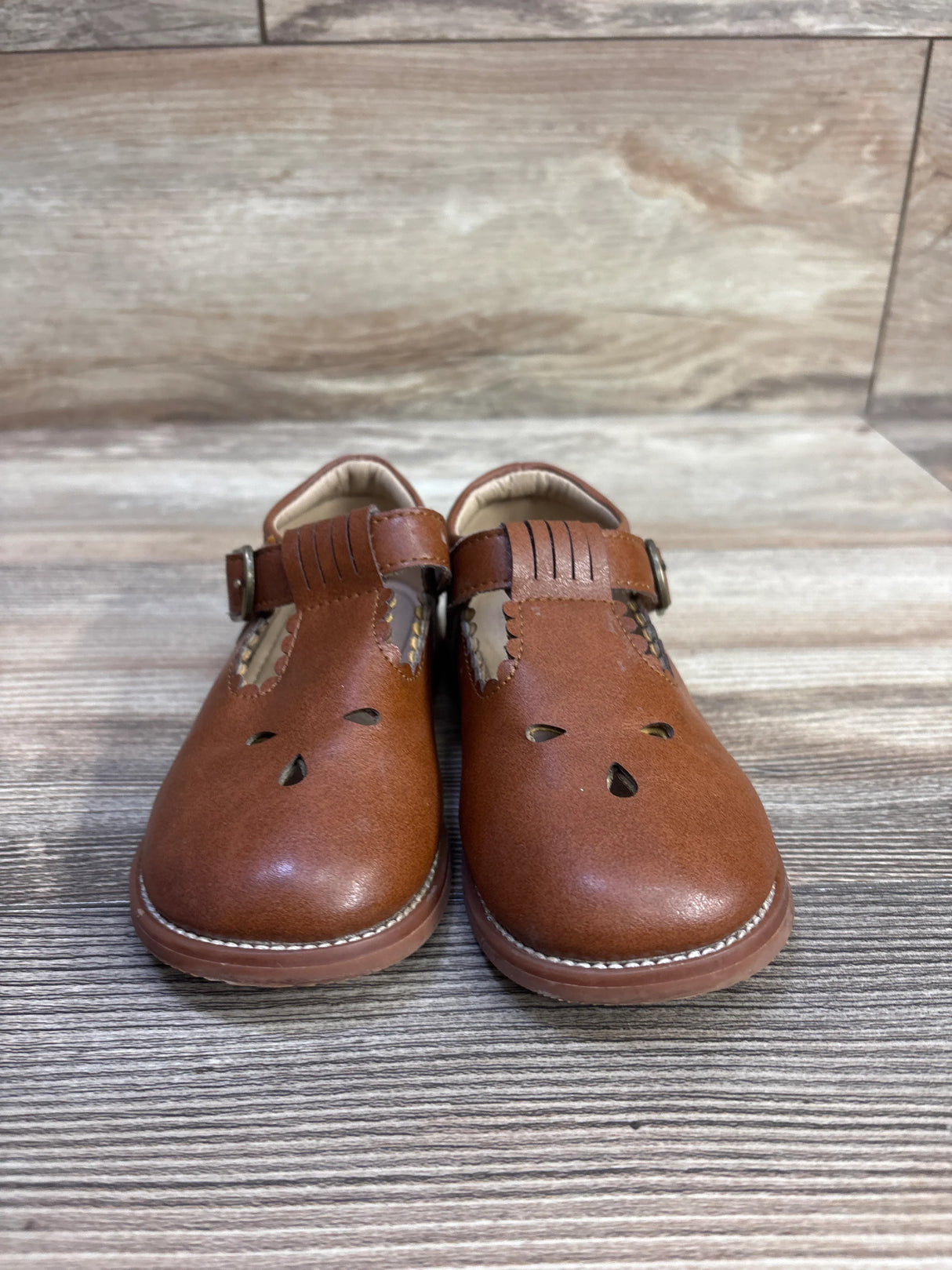 Front view of brown toddler T-strap Mary Jane shoes showing buckle strap, scalloped trim, teardrop cutouts on wood-grain surface