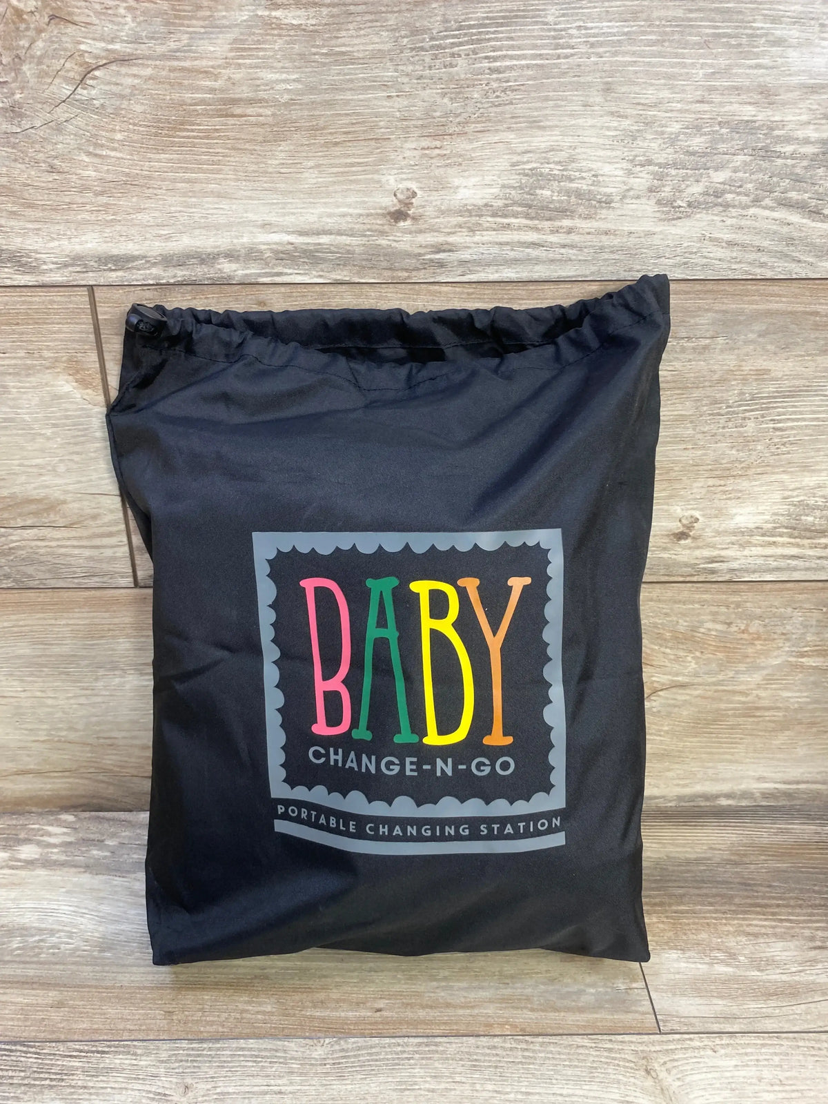 Front view of Baby Change-N-Go portable hanging changing station folded in a black case with colorful BABY logo on wood floor