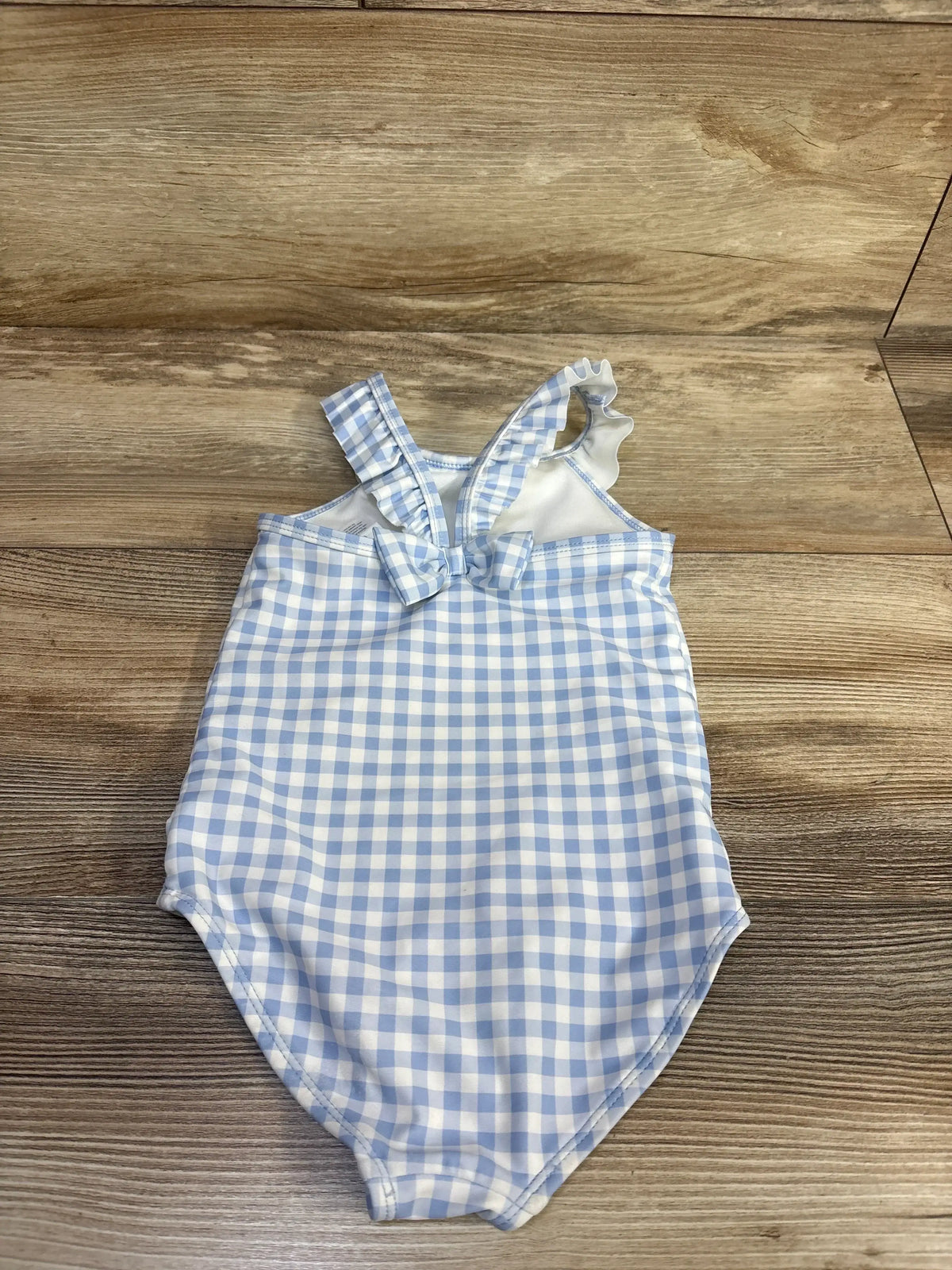Top view of toddler one-piece swimsuit with blue and white gingham, ruffle shoulder straps and front bow on wood background