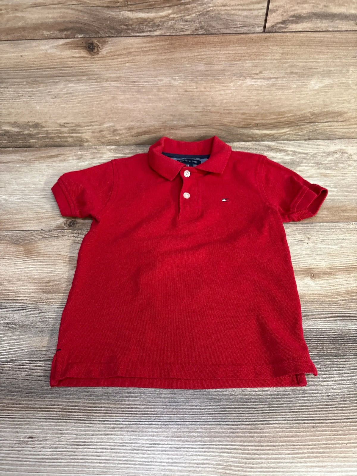 Flat lay of red Tommy Hilfiger 2T polo shirt showing two button placket and small flag logo on light wood background