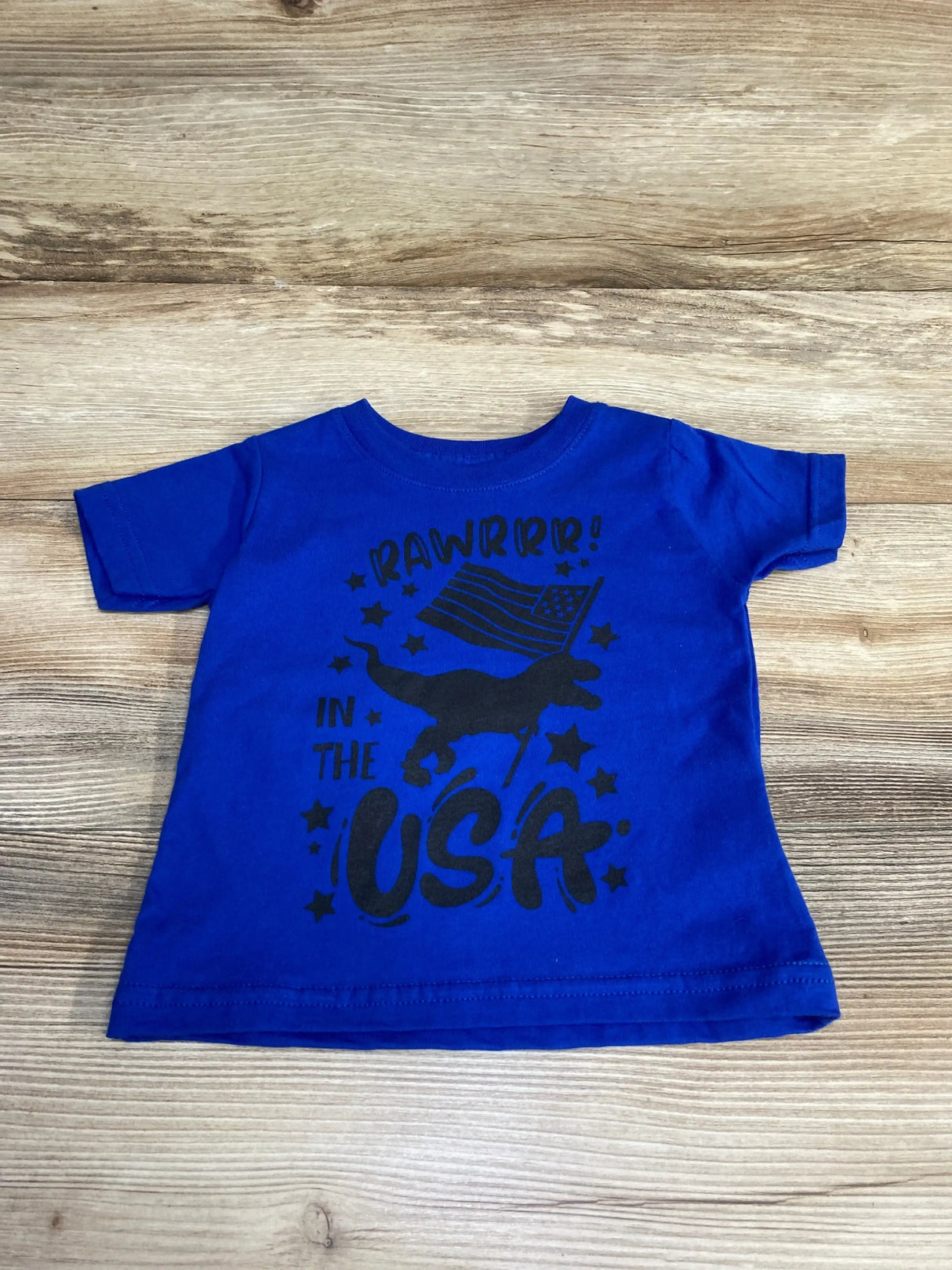 Flat lay of blue Rabbit Skins toddler T-shirt showing black Rawrrr in the USA dinosaur and American flag graphic on wood background
