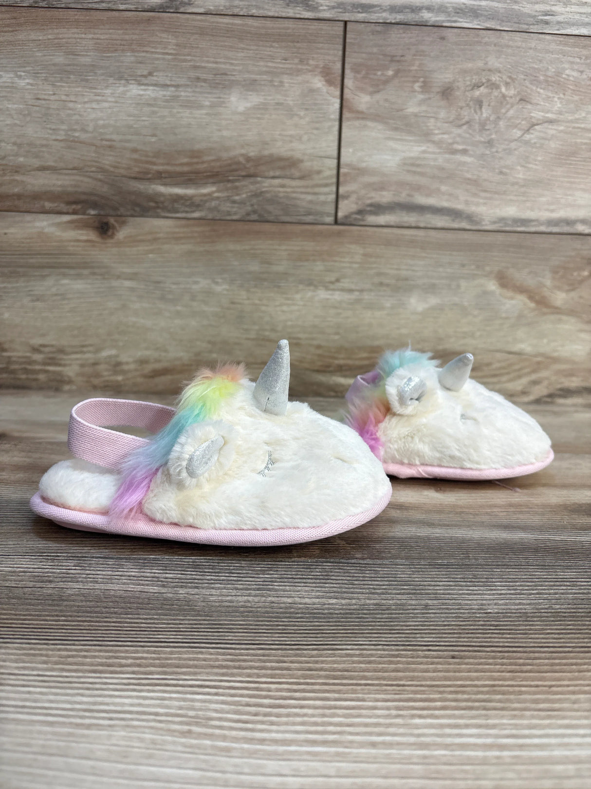Soft unicorn slippers for kids with rainbow mane, pink trim, and silver horn on wood floor