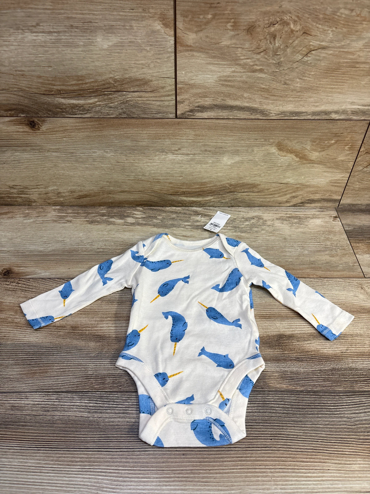 Flat lay of Old Navy ivory long-sleeve 0-3M bodysuit showcasing blue narwhal print and snap crotch on wood plank background