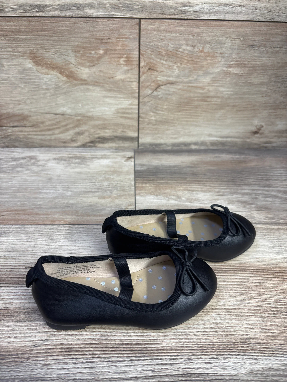 Side view of Cat & Jack Nora black slip-on ballet flats showing strap and bow on light wood background