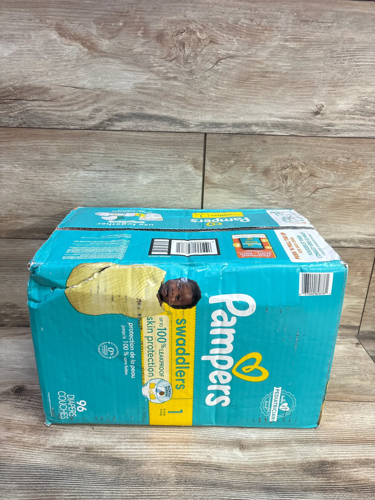 Front view of Pampers Swaddlers Size 1 diaper package showing 96 diapers and torn packaging on a wood floor
