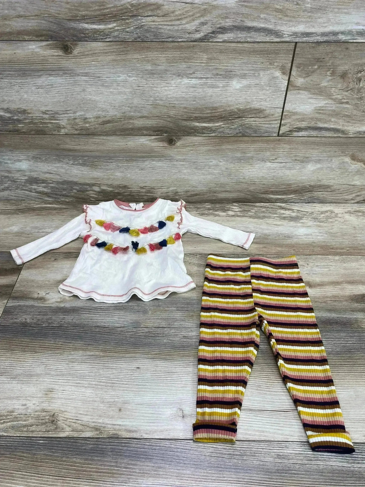 Flat lay of Mud Pie 2-piece set, size 3-6 months, cream tassel top and ribbed mustard-pink striped leggings on wood
