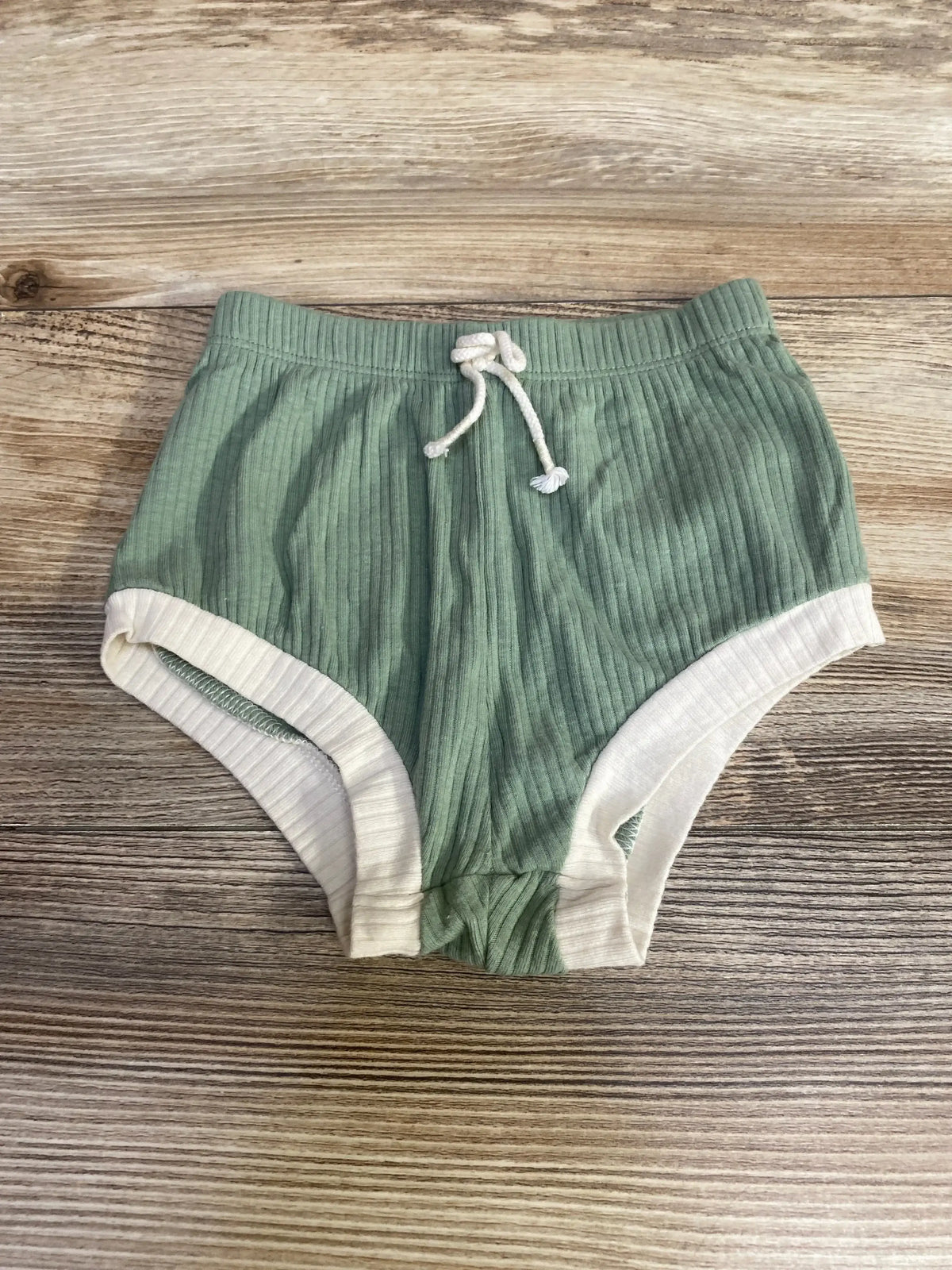 Top-down view of green ribbed baby bloomers (12 months) showcasing white leg trim and drawstring elastic waist on wood background