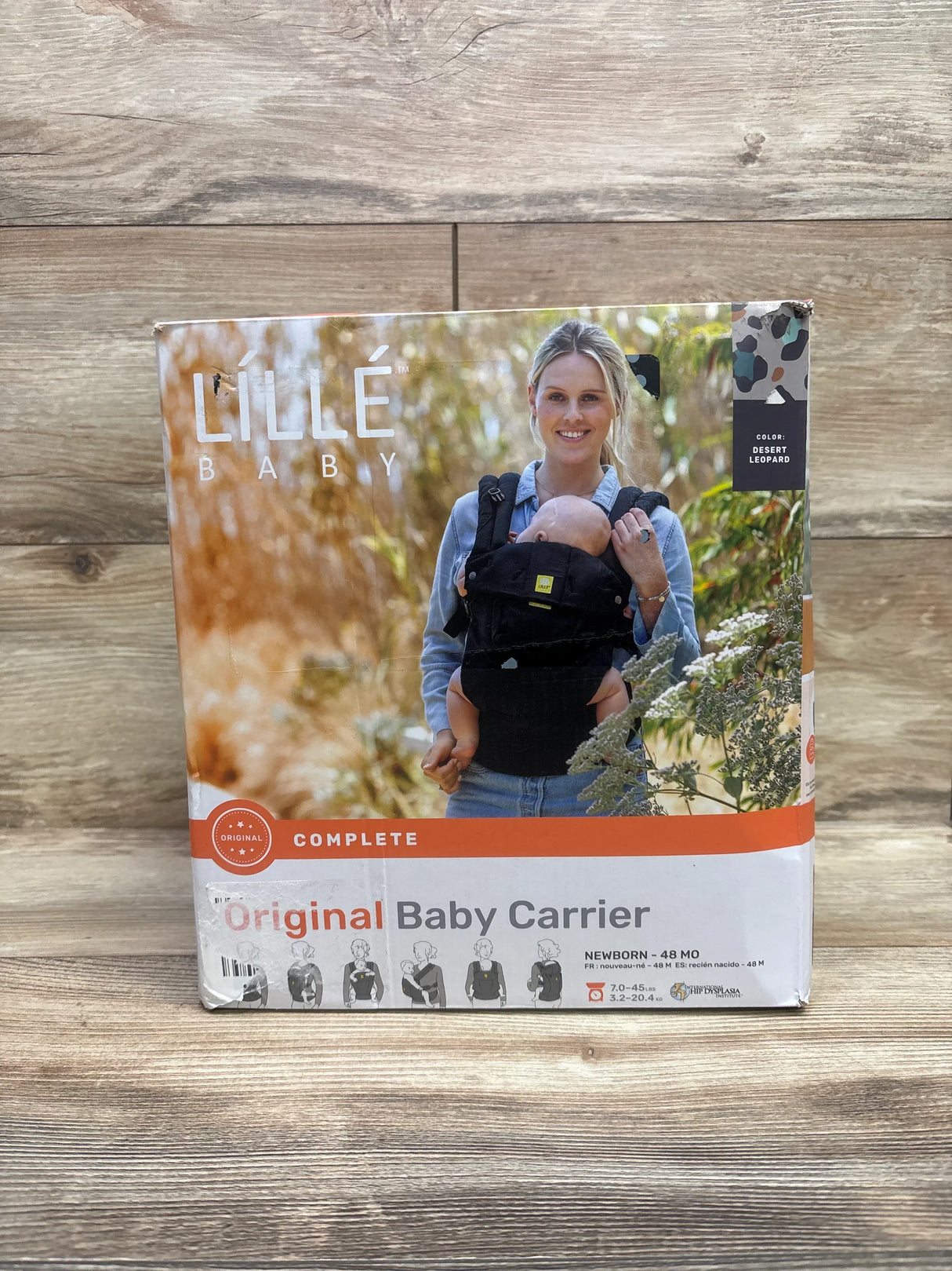 Front view of boxed LILLEbaby Complete Original baby carrier showing Desert Leopard packaging and worn box on a wooden surface