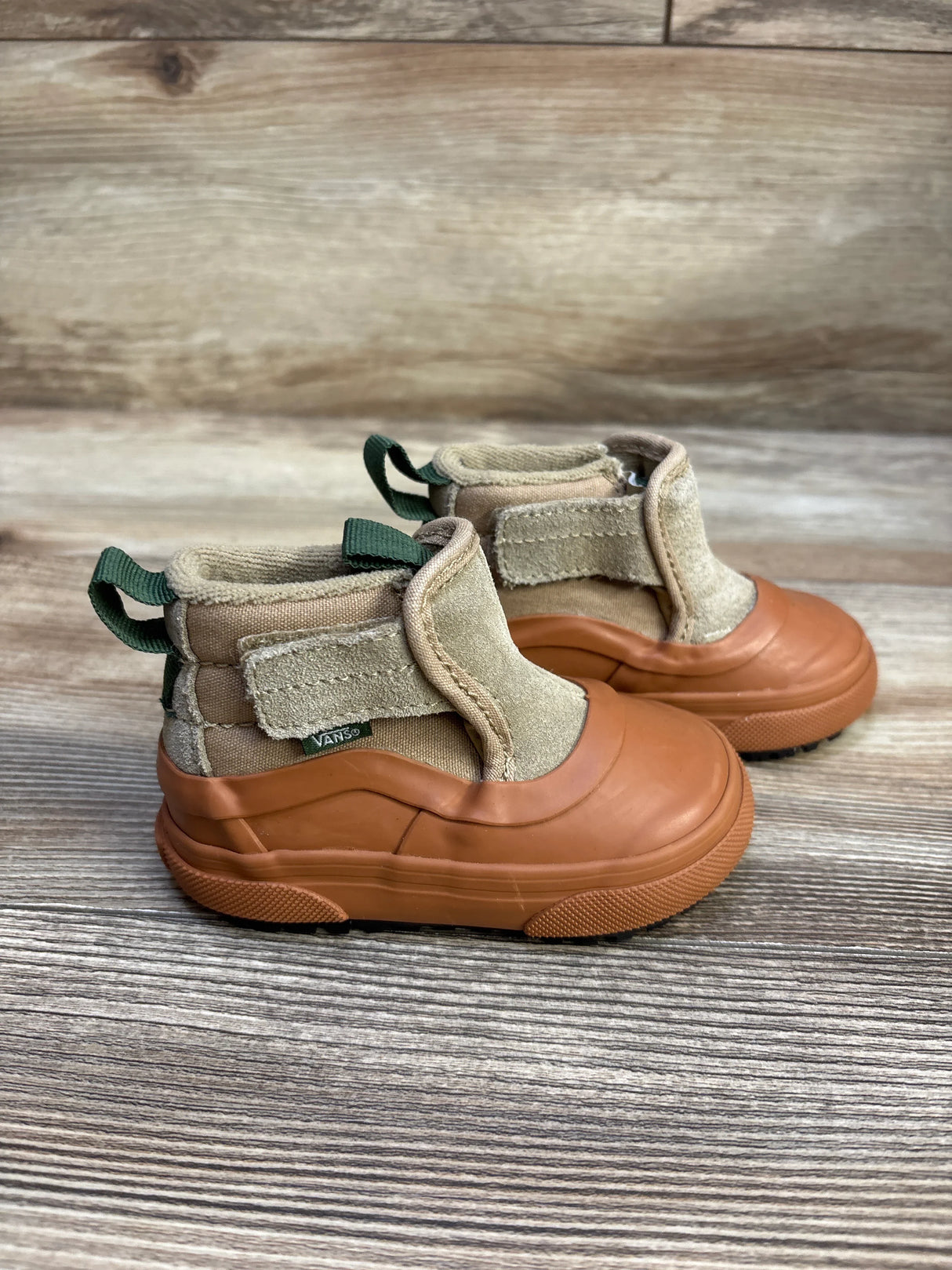 Side view of toddler Vans MTE Slip-On sneakers showing tan suede upper, brown rubber toe, green pull tab on wood background