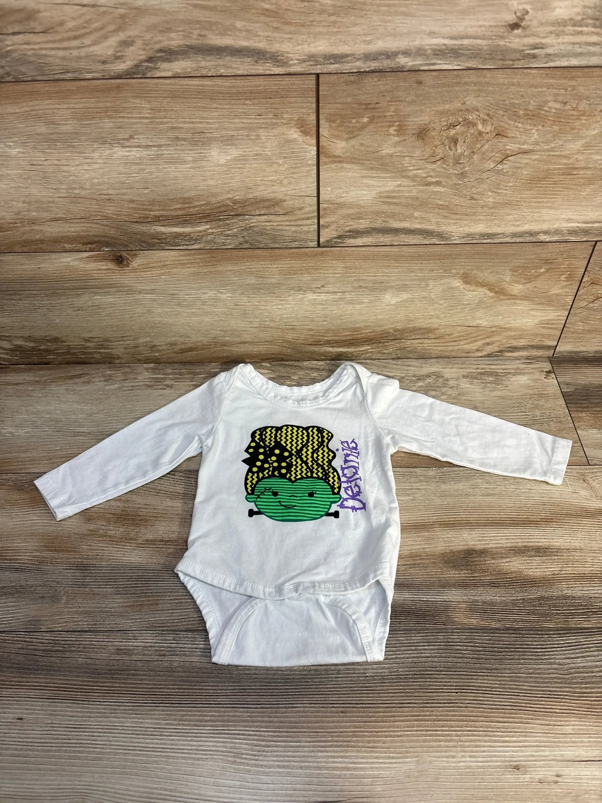 White baby onesie with green Frankenstein graphic and 'Delanie' text, laid on wood floor